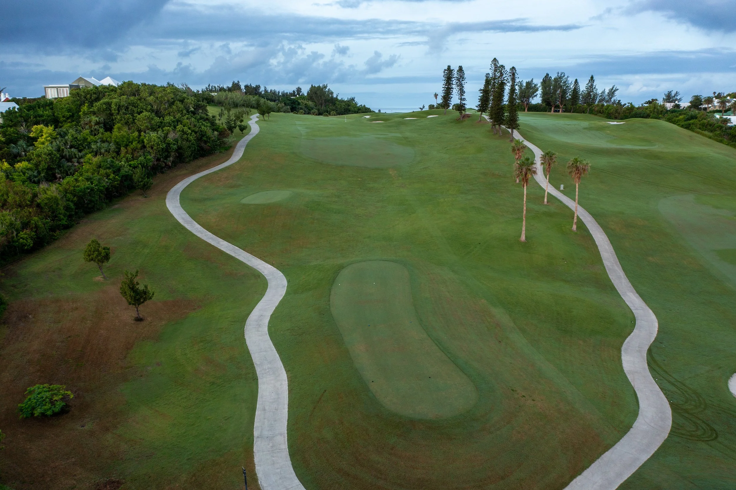 Aerial view of a golf course with winding white concrete pathways, green fairways, and several tall palm trees, under a cloudy sky.