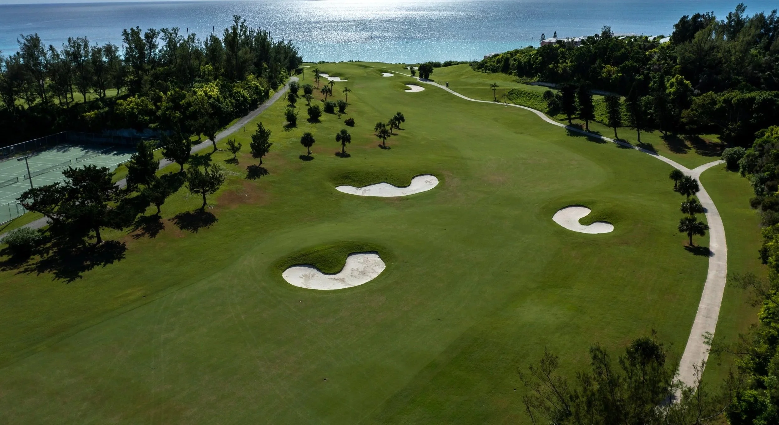 Aerial view of a golf course with sand traps, green grass, trees, and a path, overlooking a body of water.