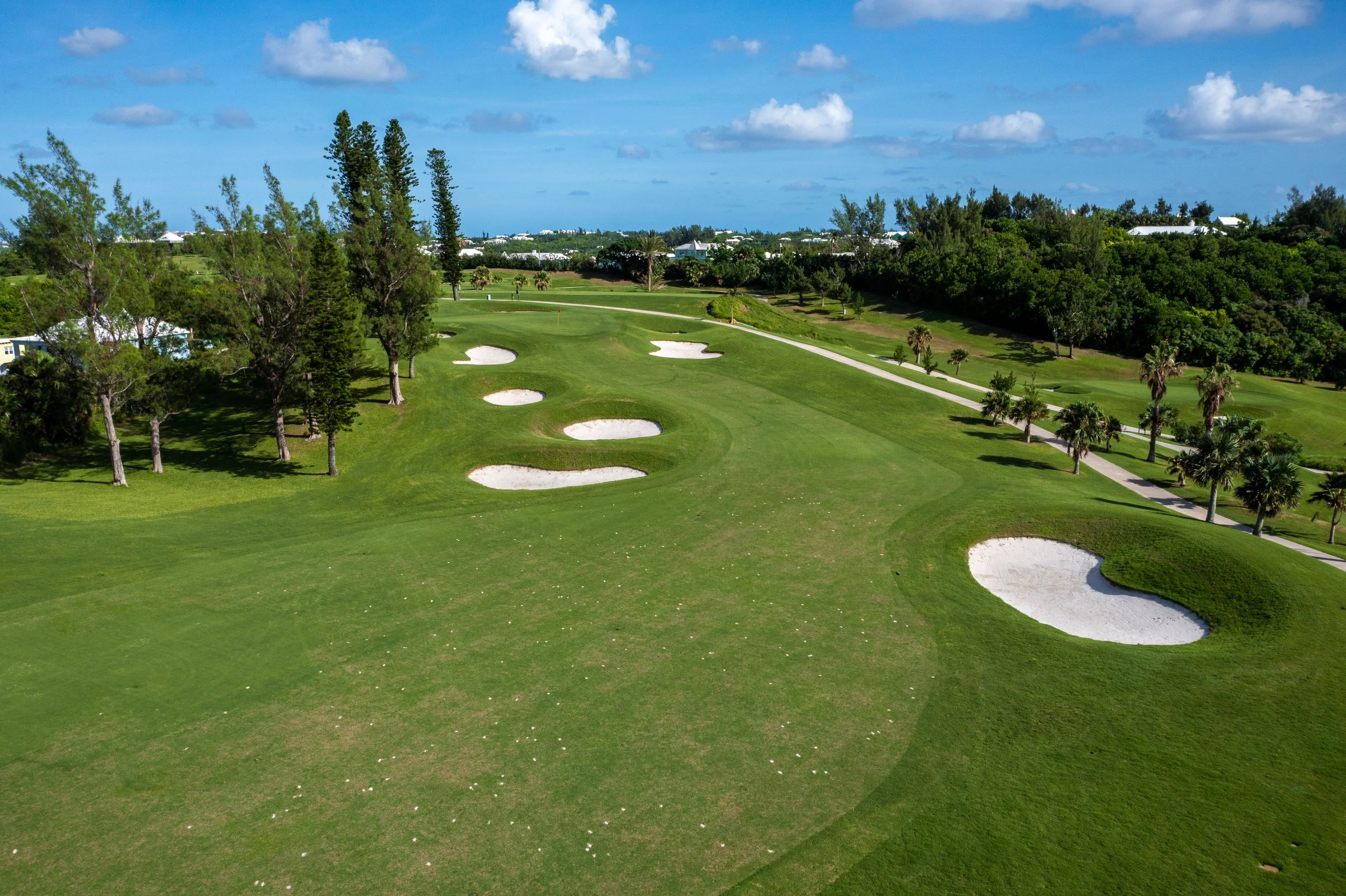 A golf course with lush green grass, sand bunkers, trees, and a clear blue sky with clouds.
