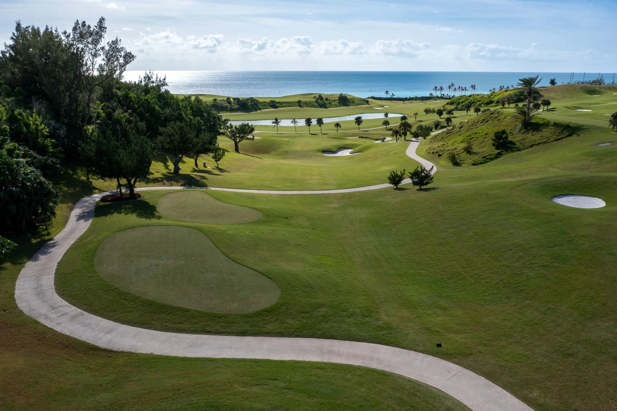 A scenic view of a golf course near the coastline with green grass, sand bunkers, and a winding concrete path. The background shows the ocean under a partly cloudy sky, with palm trees lining the horizon.