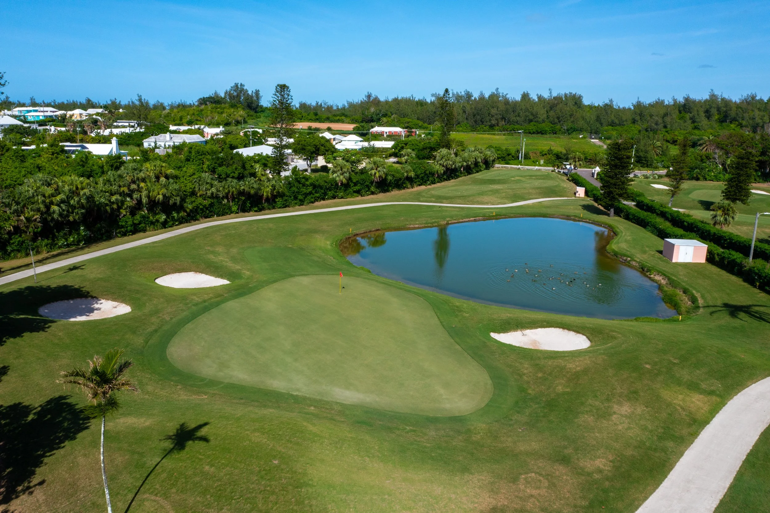 An aerial view of a golf course with a pond, sand bunkers, grassy areas, and a paved cart path. In the background, there are trees and houses with white roofs, under a blue sky.
