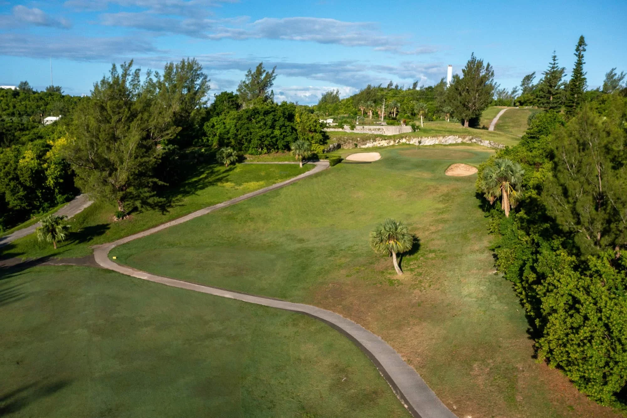 A scenic view of a golf course with lush green grass, sand traps, and a winding pathway, surrounded by tall trees and a partly cloudy blue sky.