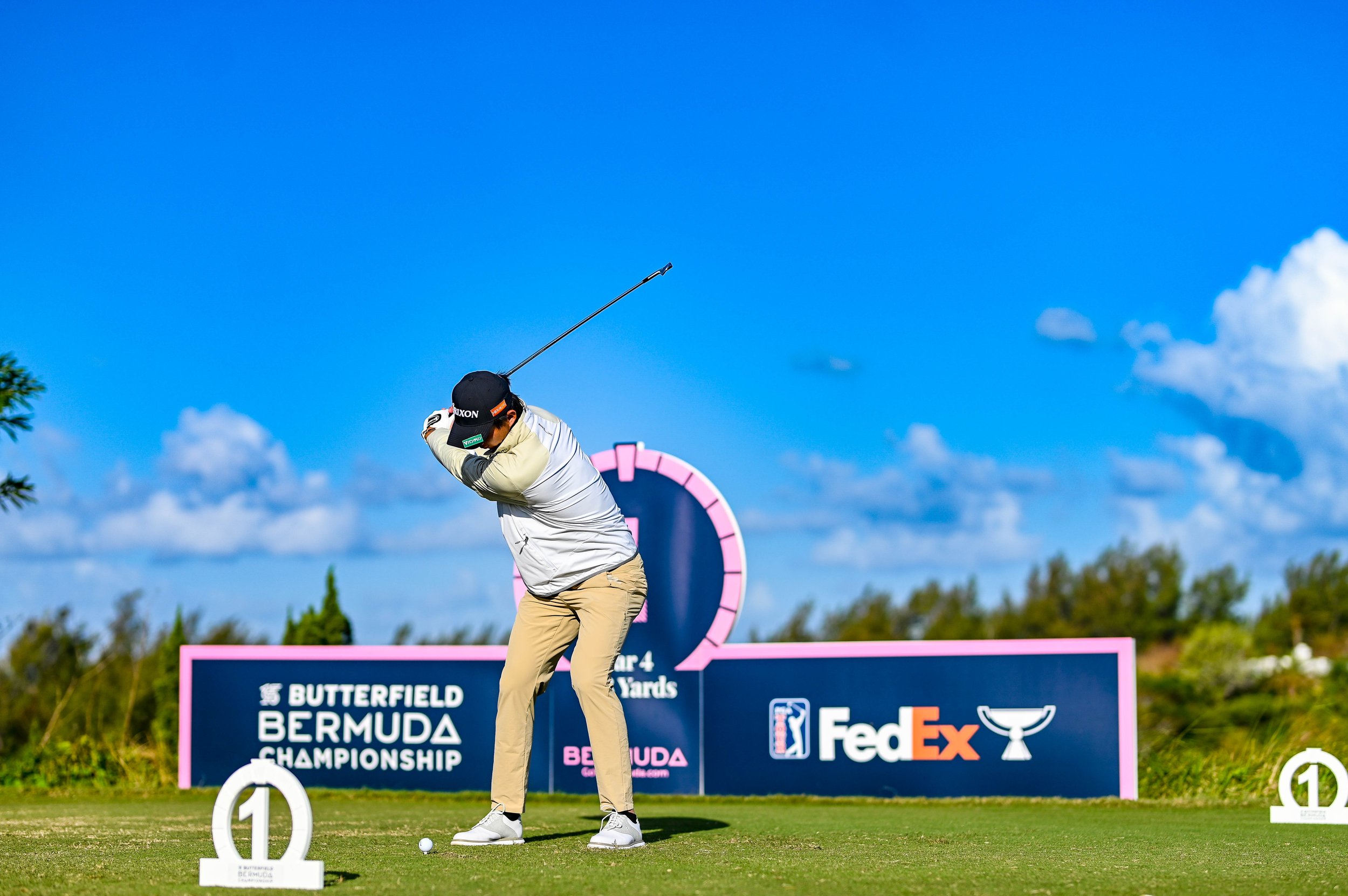 A golfer taking a shot during the Bermuda Championship, with signage displaying sponsor logos and a blue sky with clouds in the background.