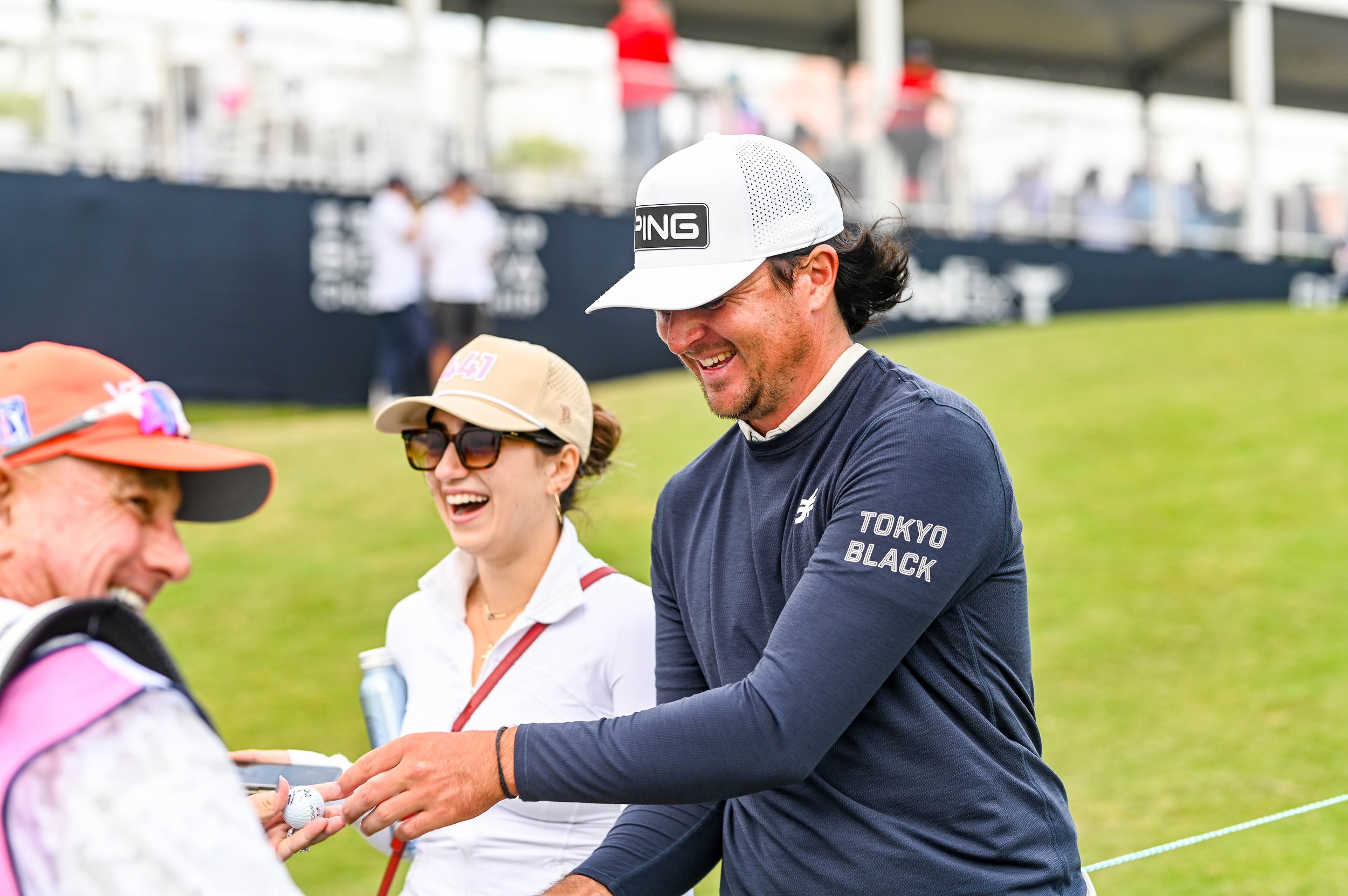 People smiling and chatting at an outdoor event, with a man in a white cap with the PING logo and a woman wearing sunglasses and a beige cap, on a grassy area with a blurred background of other individuals and event structures.