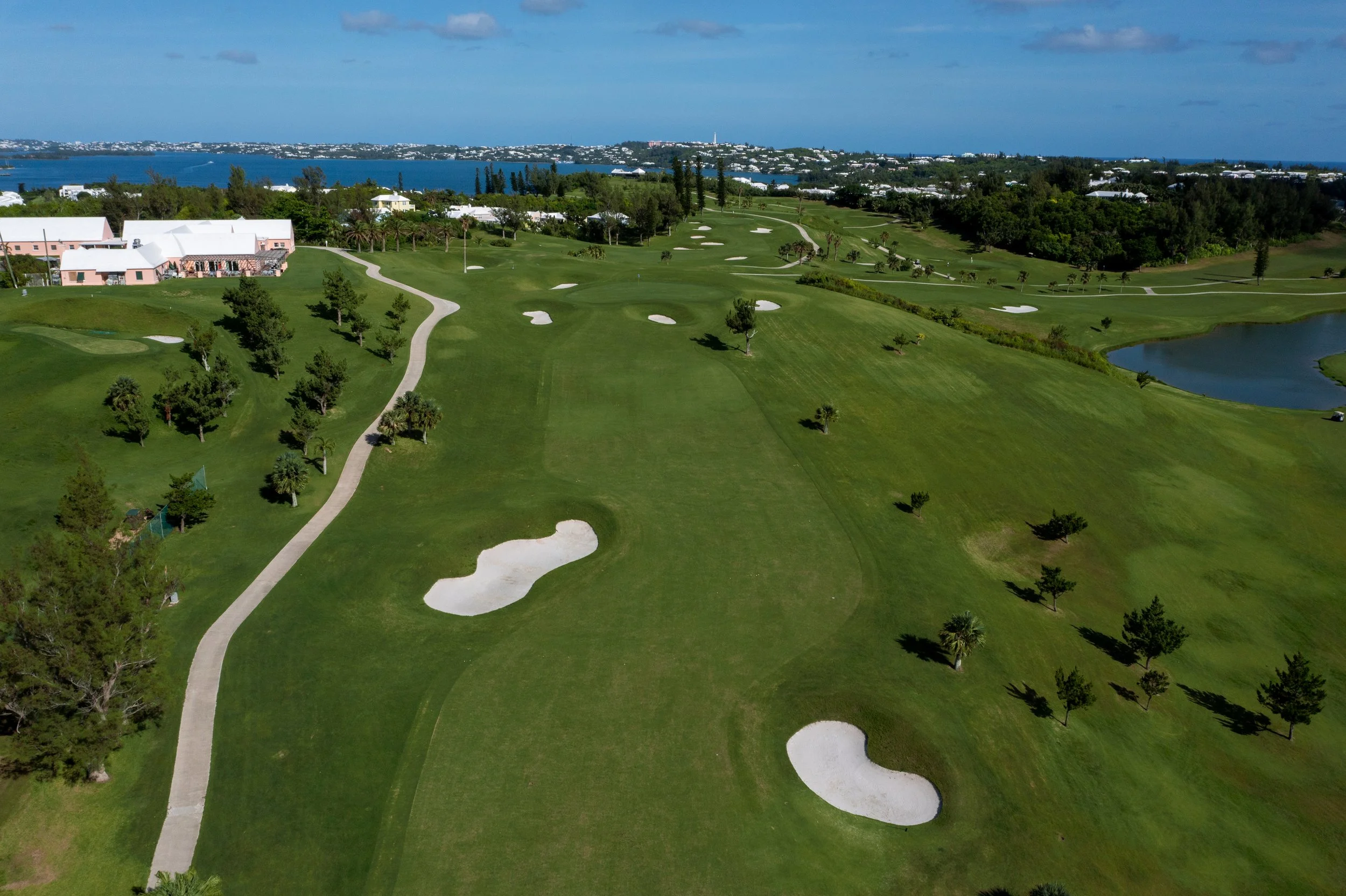Aerial view of a golf course with green fairways, sand traps, and trees, near a body of water and cityscape in the background.