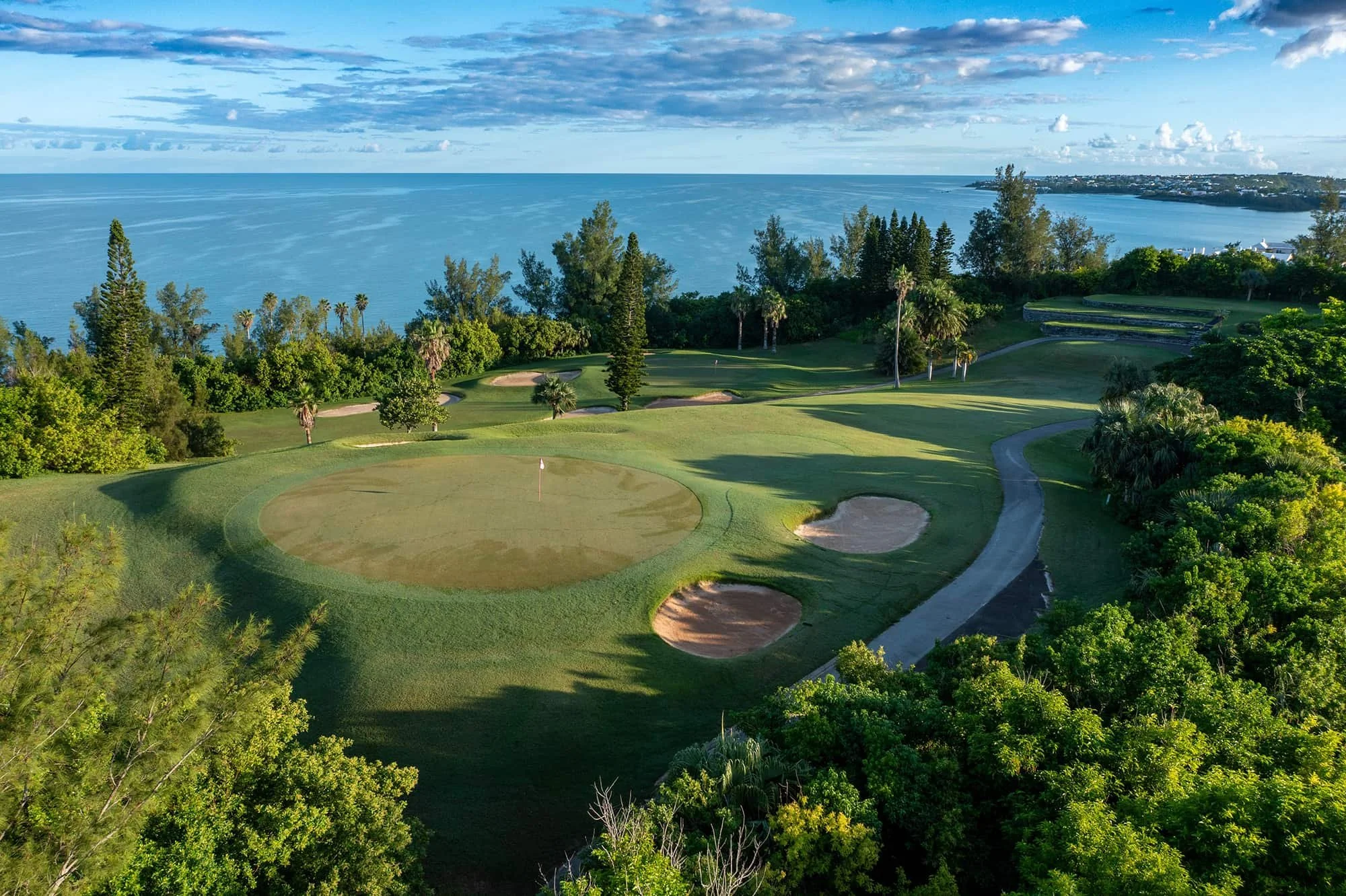 An aerial view of a golf course by the ocean, featuring sand traps, green fairways, and a backdrop of water with distant land and boats under a partly cloudy sky.