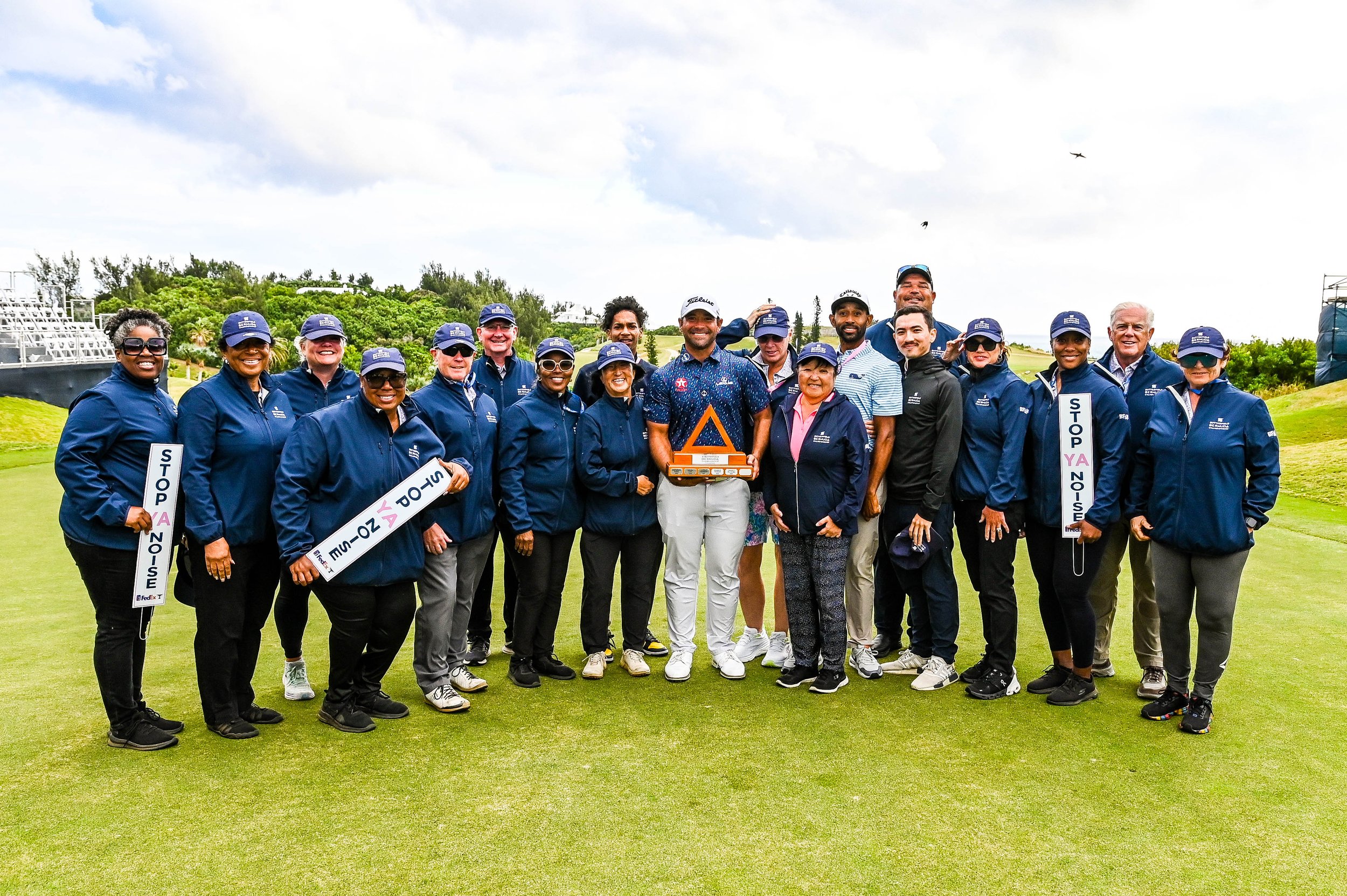 Group of people on a golf course, celebrating with trophies and signs promoting stopping noise. They are wearing golf caps and jackets, smiling, with greenery and a cloudy sky in the background.