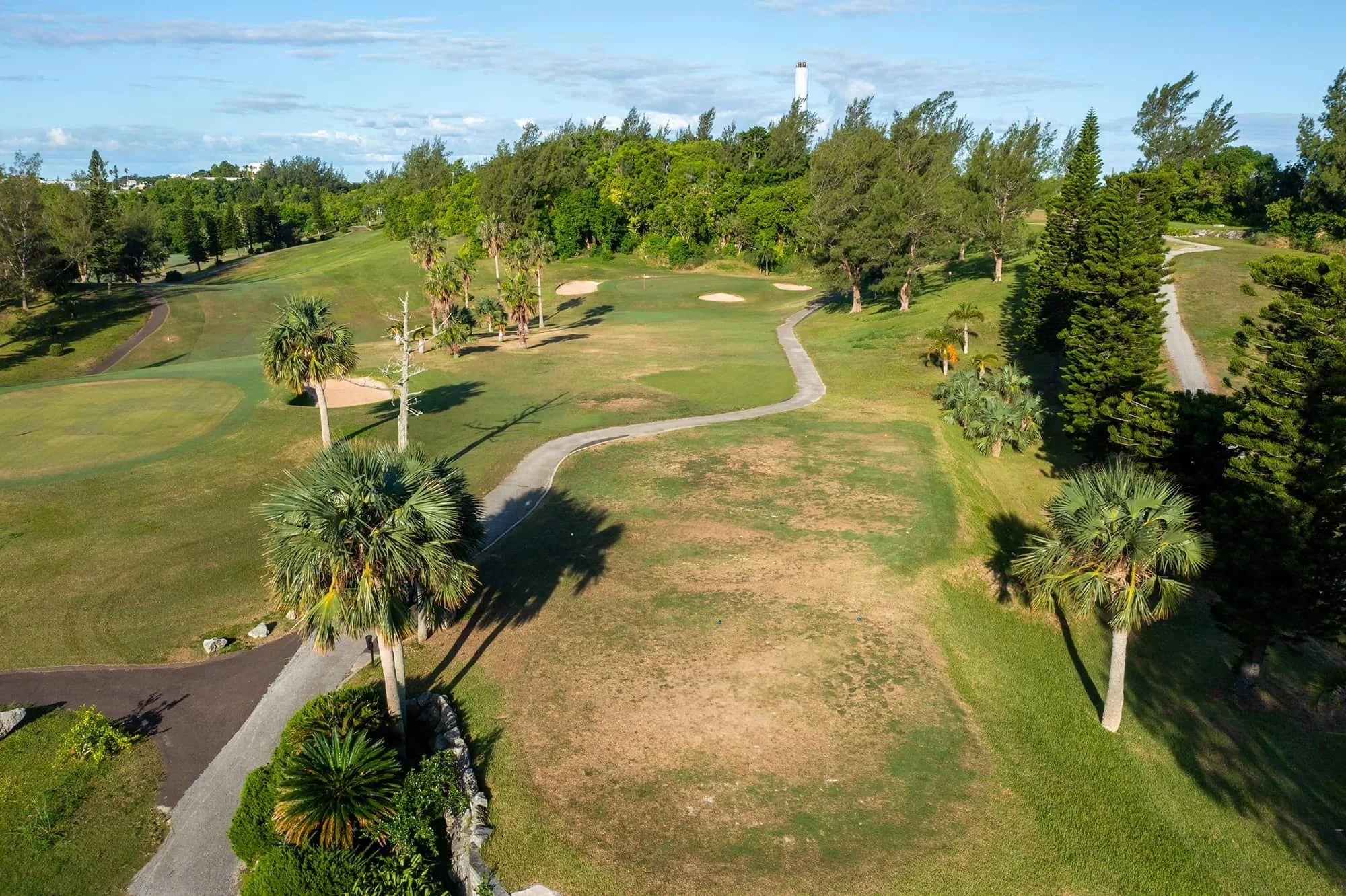 Aerial view of a golf course with green fairways, sand bunkers, and tall trees under a partly cloudy sky.