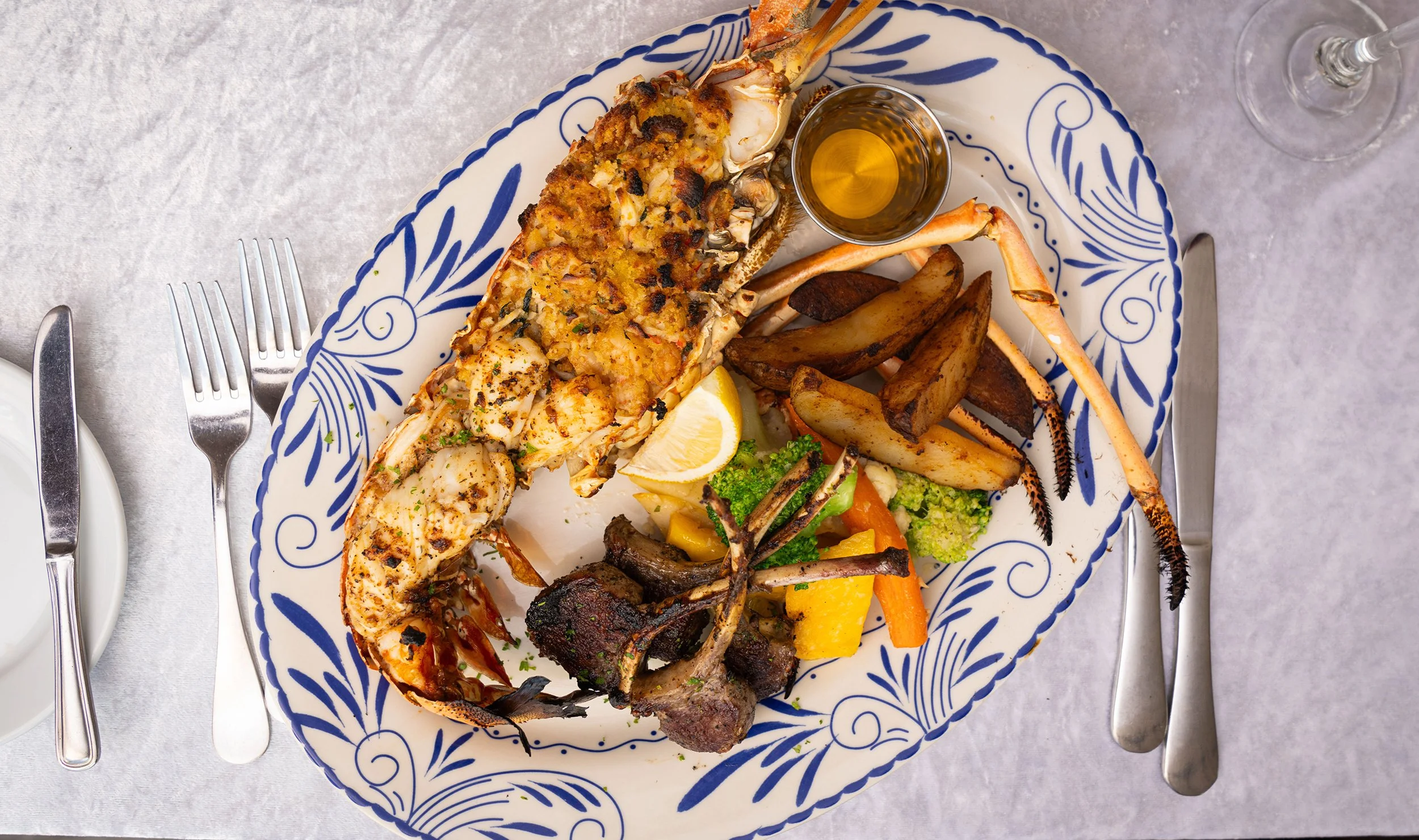 A seafood feast on a decorative blue and white plate featuring lobster tail, potatoes, vegetables, and melted cheese, accompanied by a small shot of golden sauce and served with a glass of white wine.