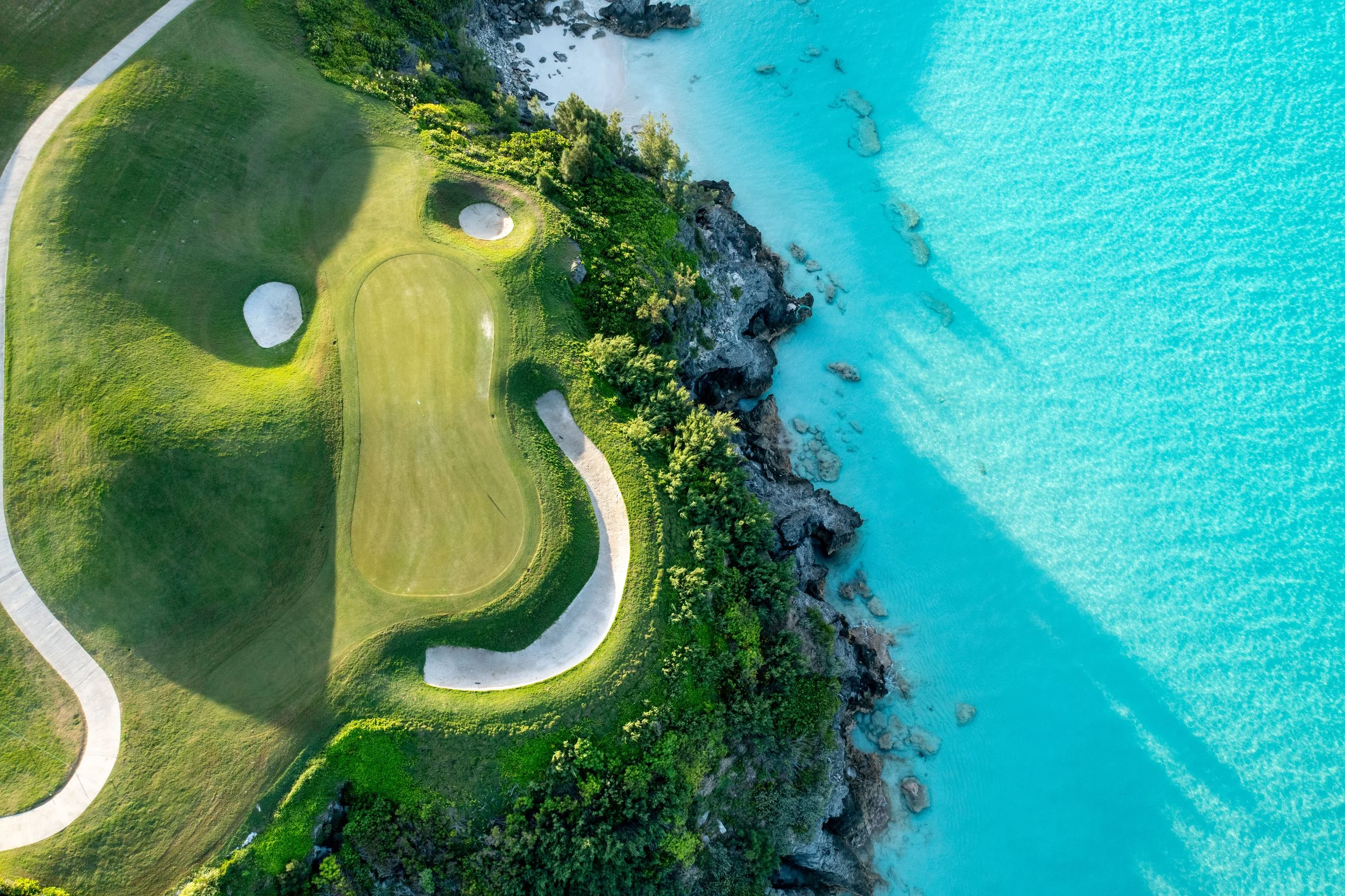 Aerial view of a golf course situated on a cliffside next to a turquoise body of water, with greenery and pathways surrounding the course.
