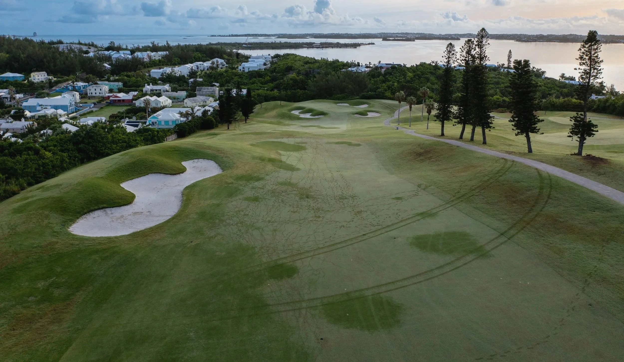 Aerial view of a golf course near water with trees, sand traps, and residential houses in the background.