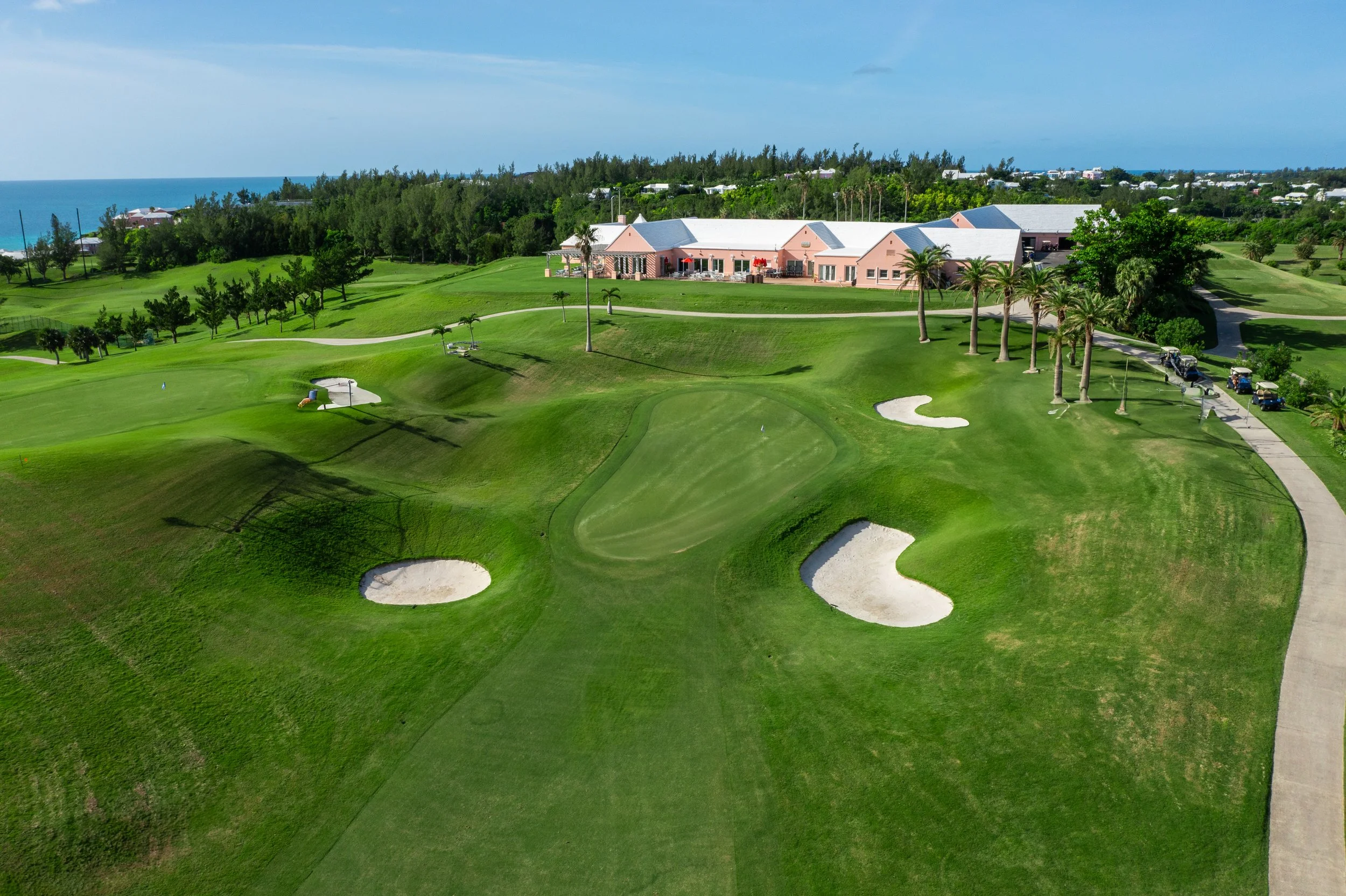 Aerial view of a golf course with green fairways, sand bunkers, palm trees, and a clubhouse overlooking the ocean in the background.