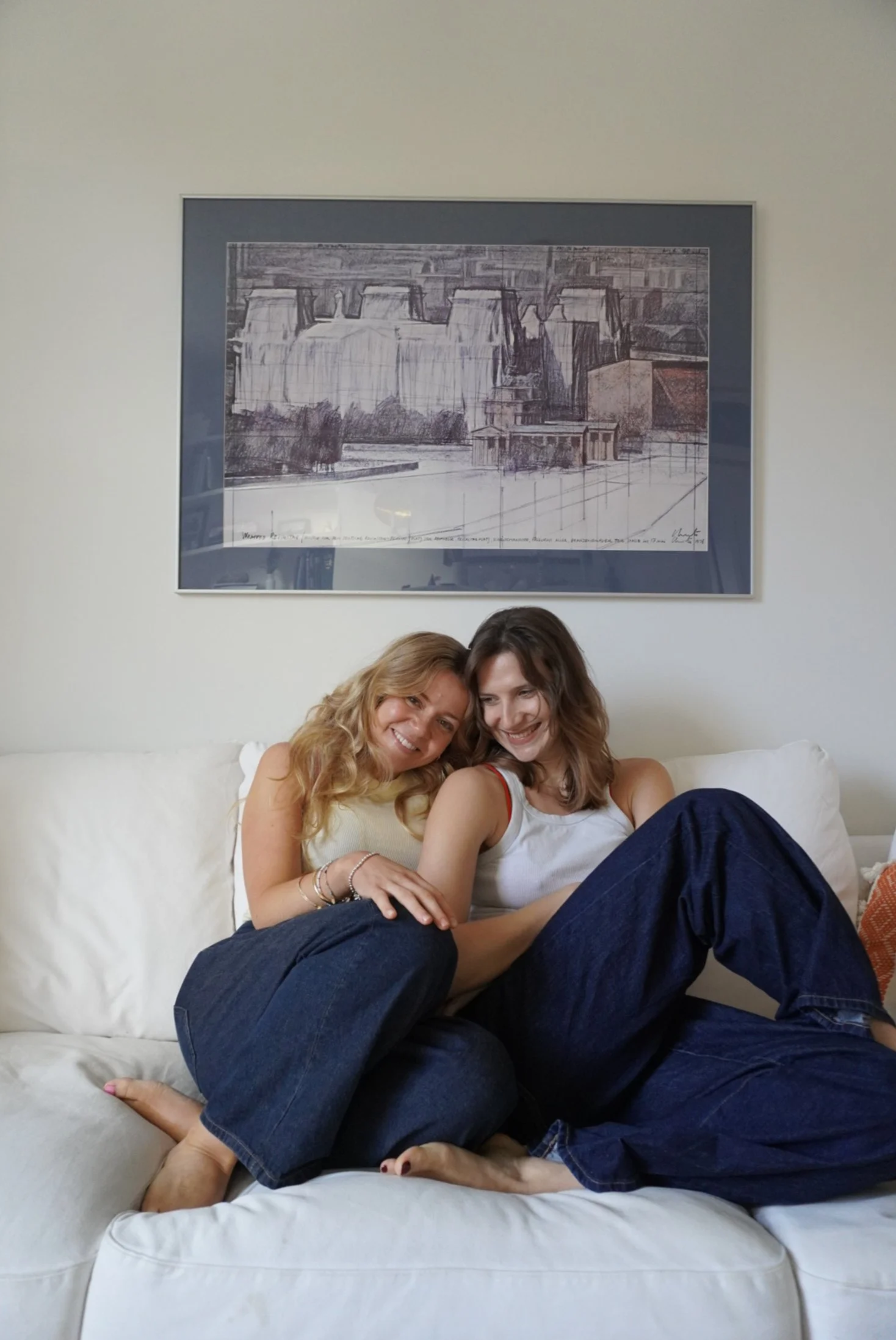 Two women sitting on a white couch, smiling and hugging each other, with a framed picture of a nuclear power plant in the background.
