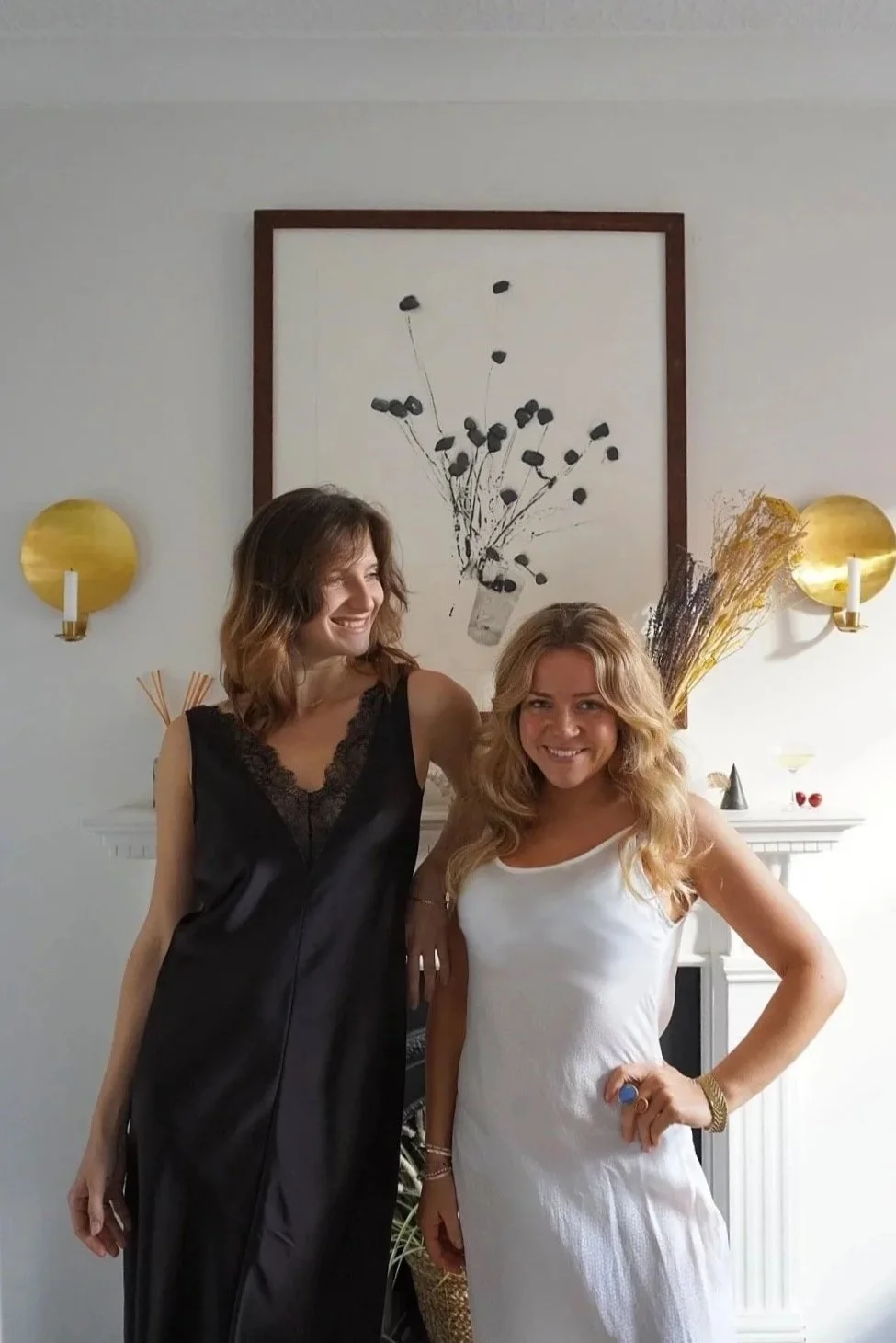 Two women smiling and standing close together indoors, one in a black dress and the other in a white dress, with a decorated mantel and wall art behind them.