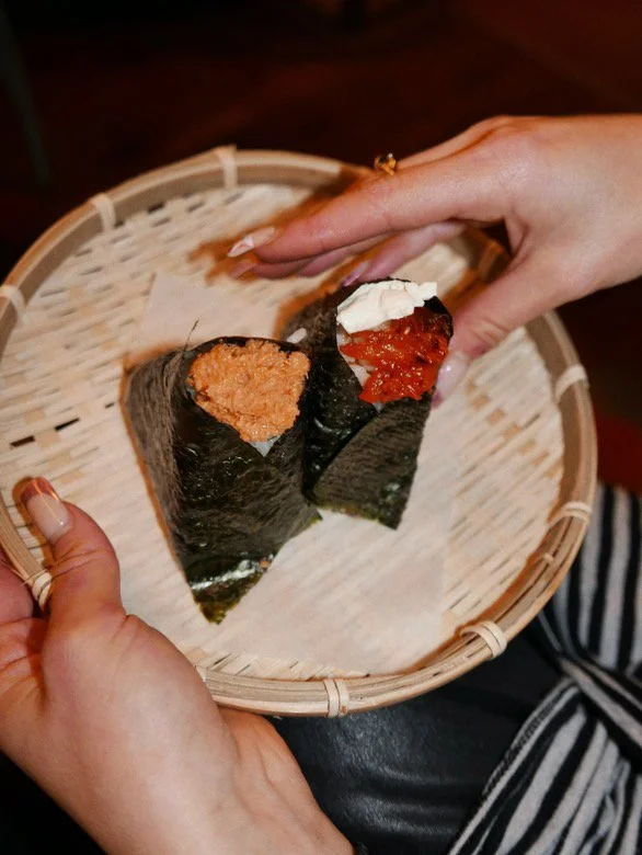 Close-up of a person holding two sushi hand rolls with seaweed wrappers on a woven basket.
