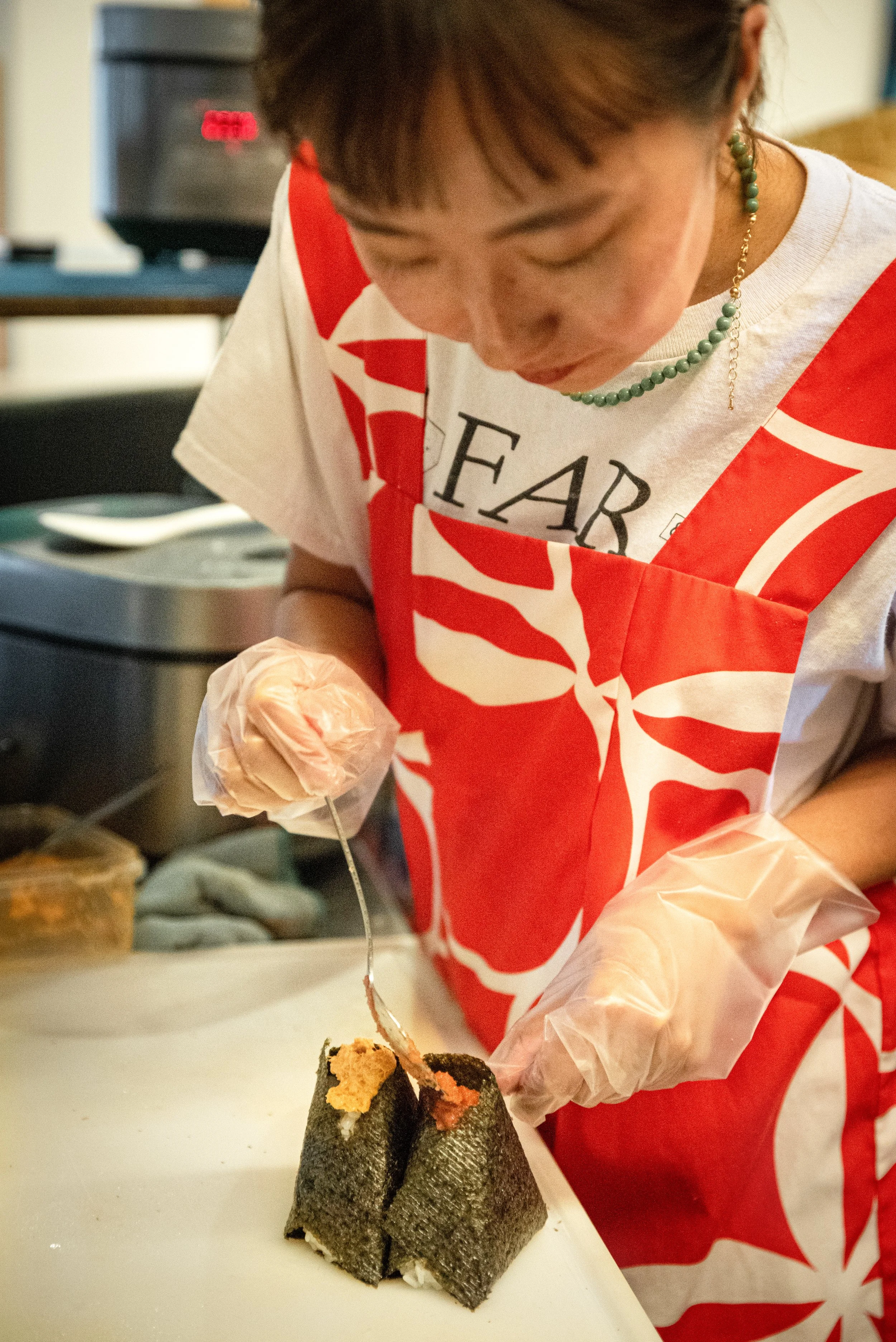 A woman wearing gloves, a red and white apron, and earrings, is preparing sushi with seaweed and rice, using a metal tool.