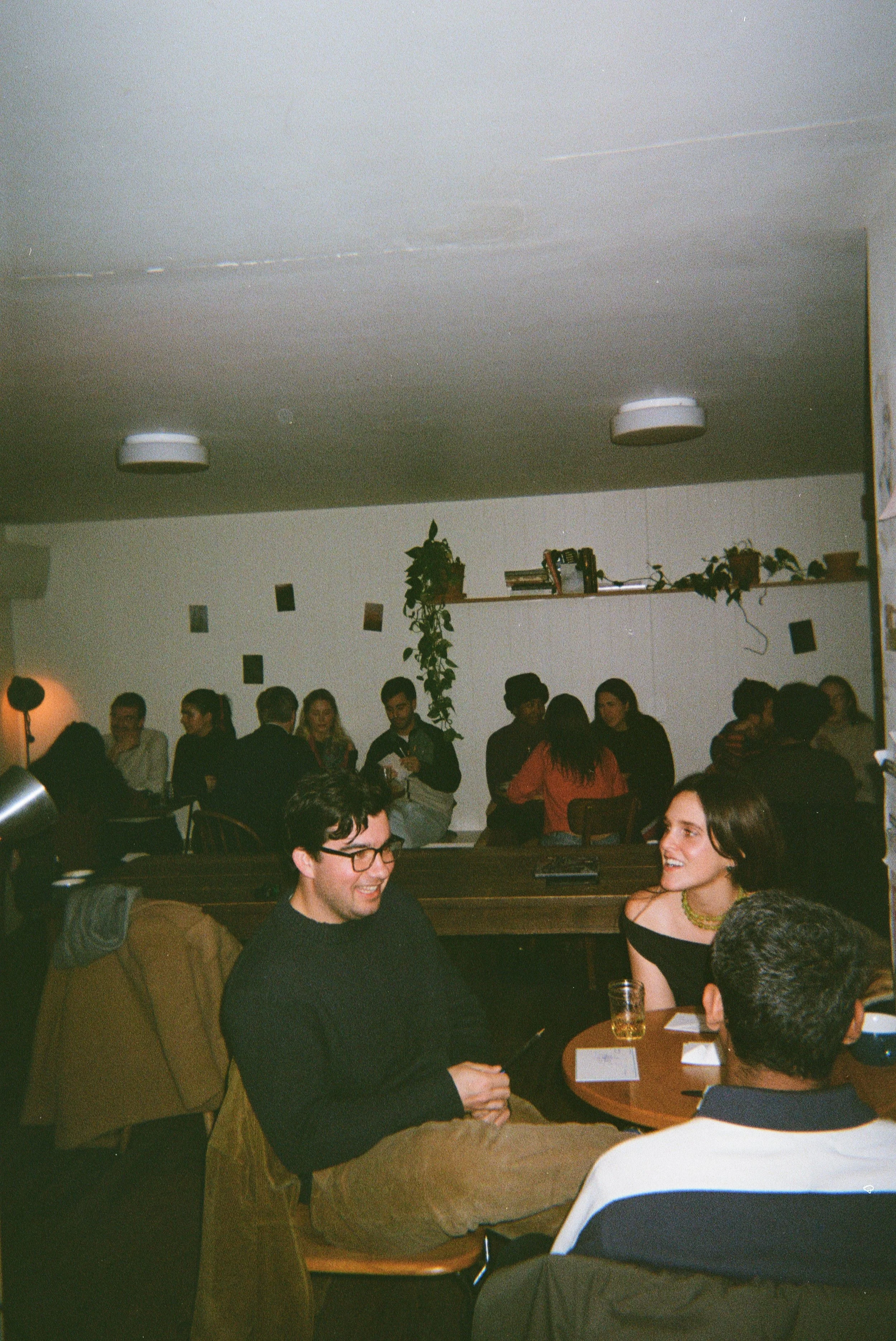 People socializing and sitting at tables in a dimly lit indoor space, possibly a bar or restaurant, with some smiling and engaging in conversation.