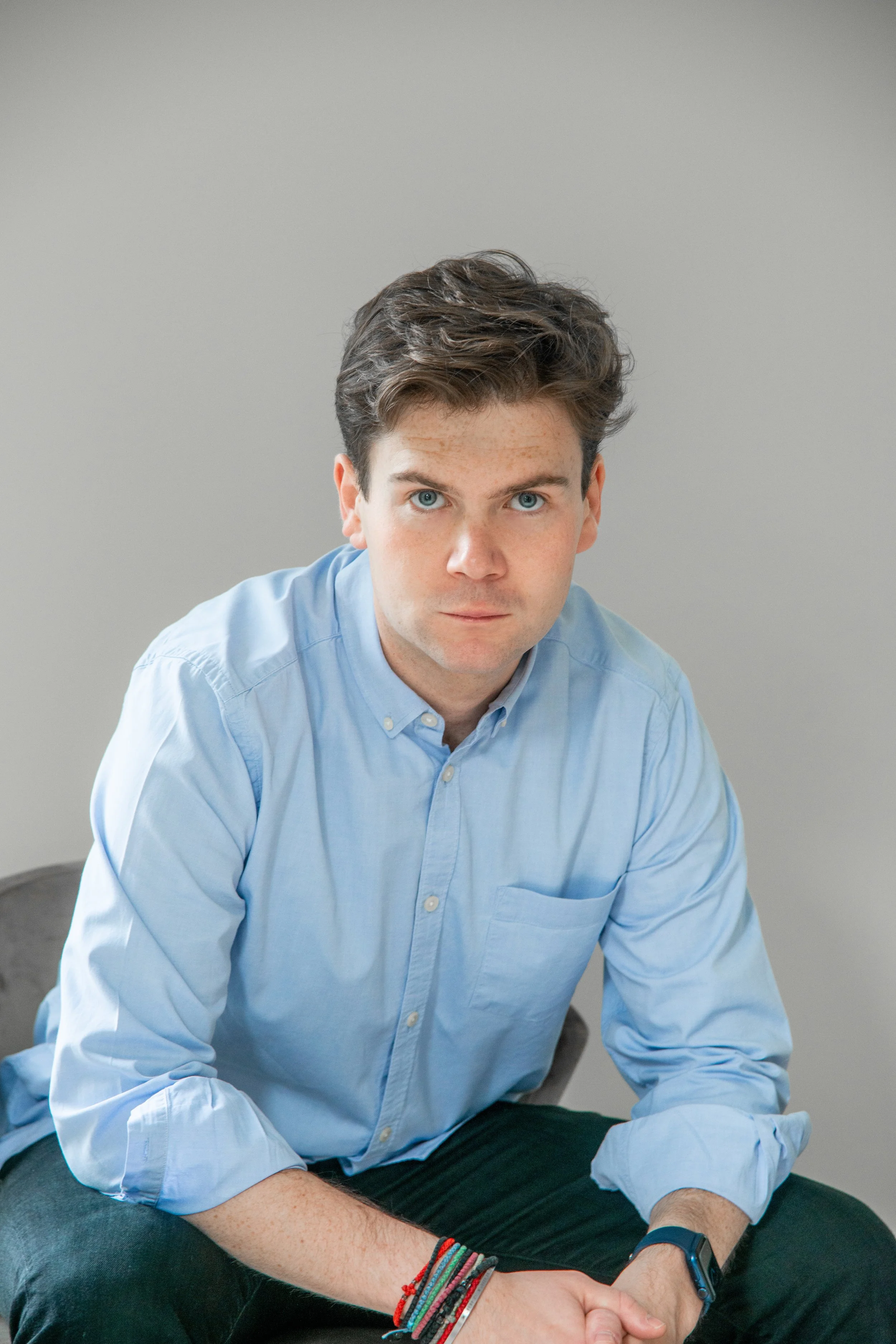 Young man with blue eyes, wearing a light blue button-up shirt, sitting on a chair with a neutral background.