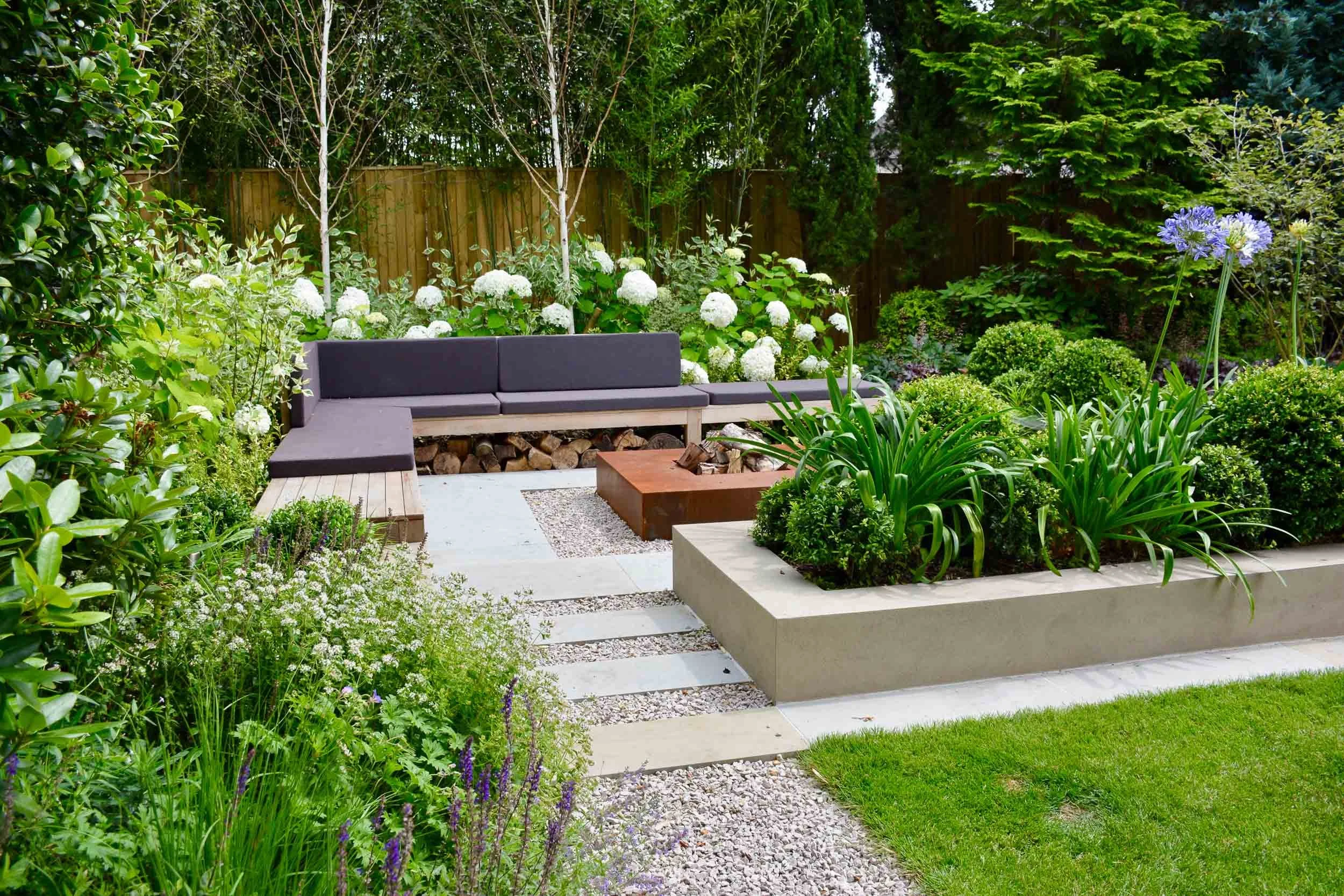 A backyard garden with a modern L-shaped patio seating area with dark cushions, surrounded by various green plants and flowering bushes, wood fire pit, and a wooden fence in the background.