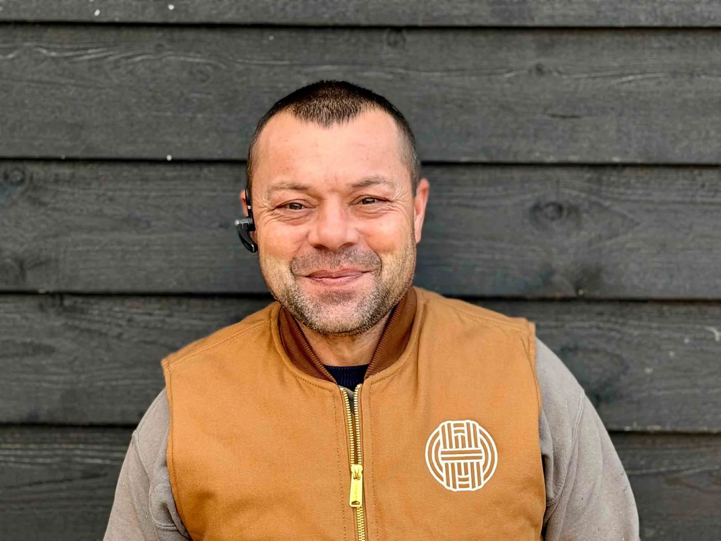 Portrait of a smiling man with short dark hair wearing a brown gilet with a circular logo against a black wooden wall