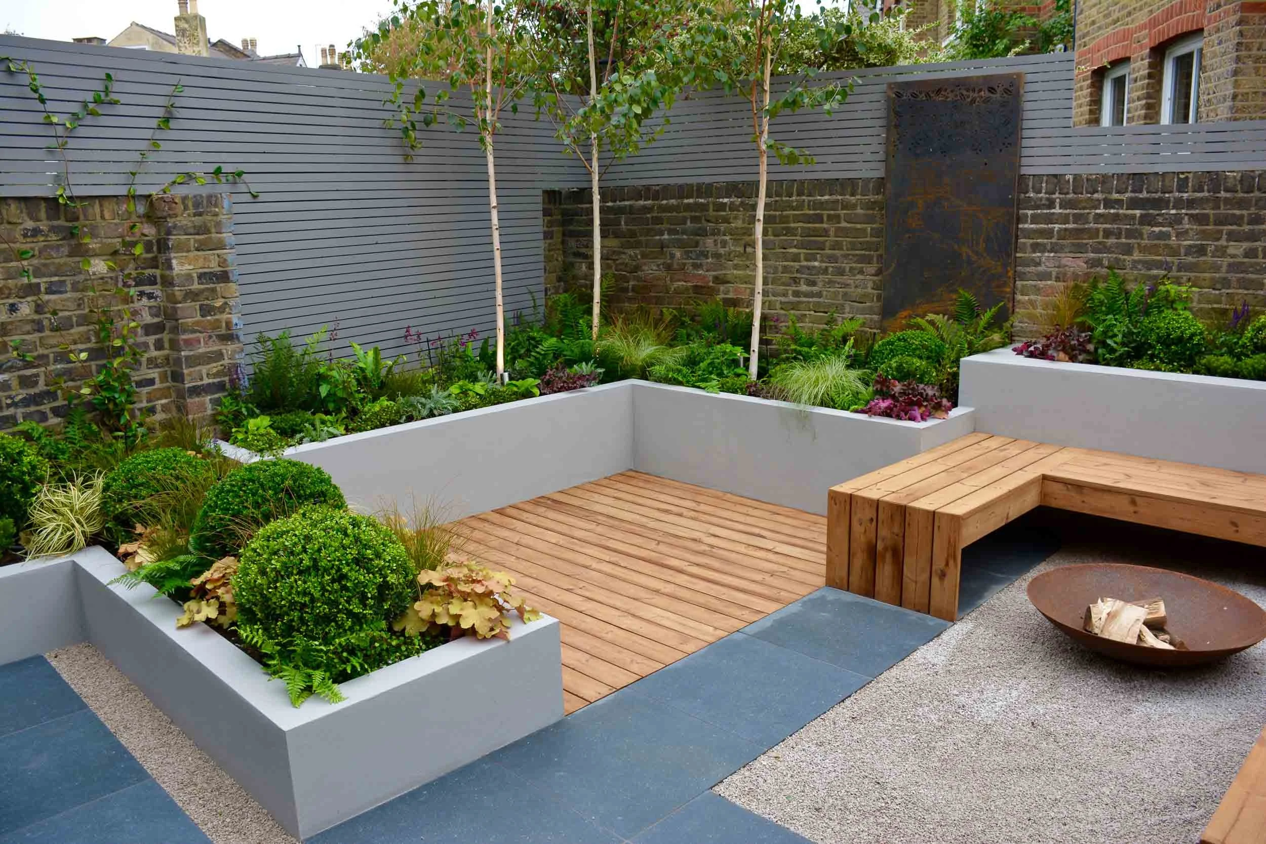 Modern backyard patio with wooden decking, built-in wooden bench, fire pit, and raised garden beds with lush green plants and small trees, enclosed by a brick and modern fence.