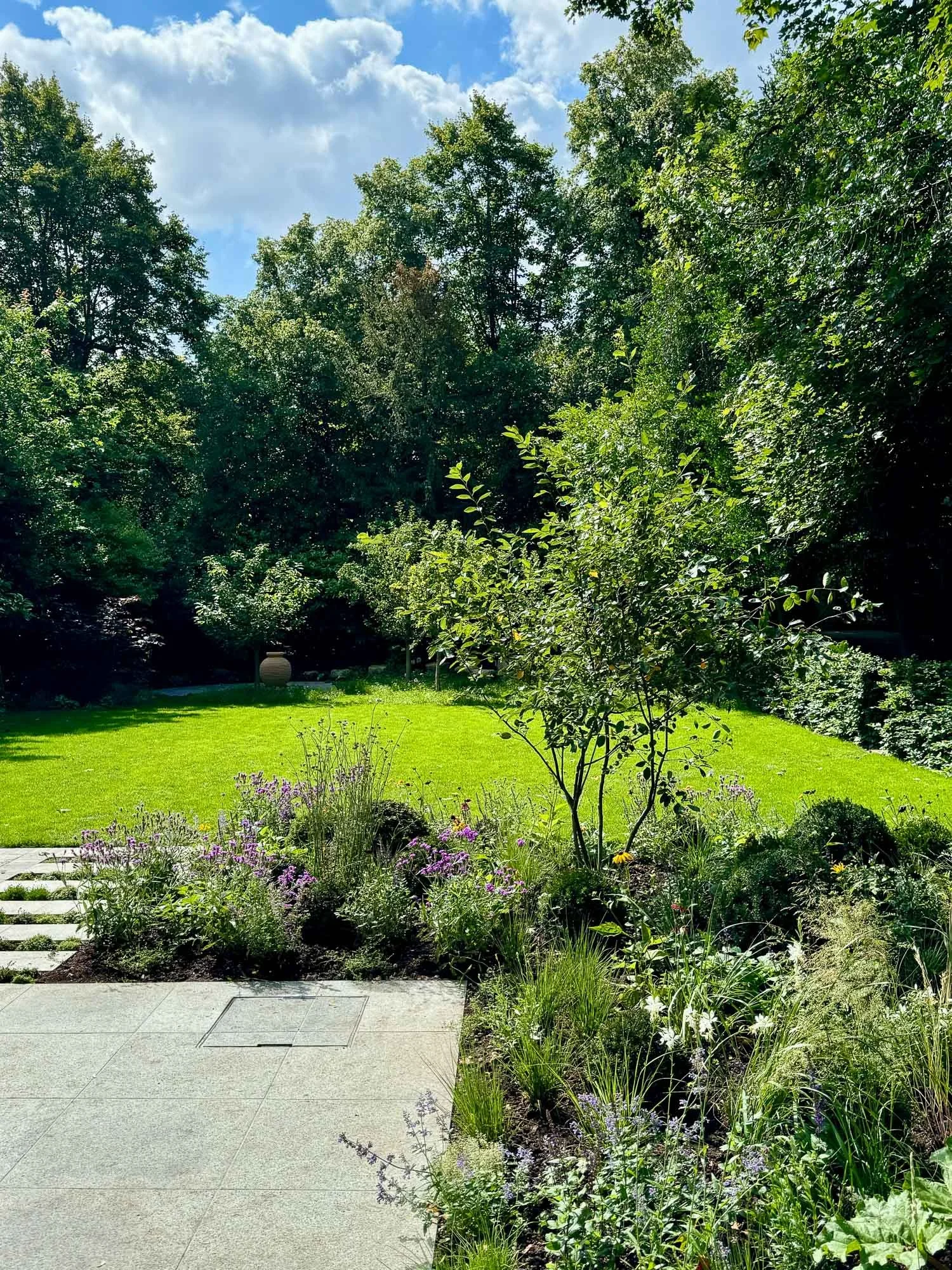 A lush backyard garden with a green lawn, surrounded by trees and shrubs, with a paved patio in the foreground.