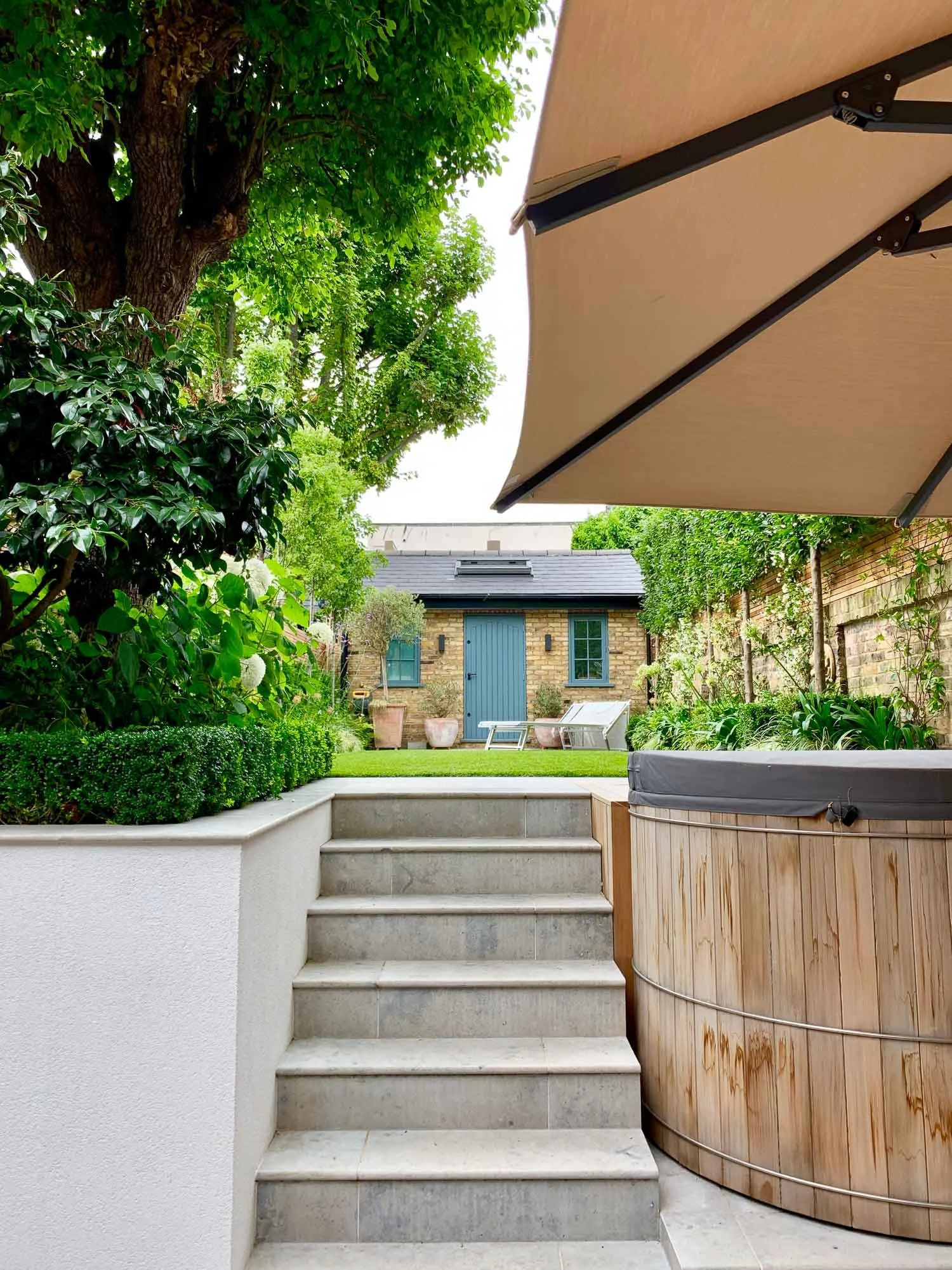 View of a backyard patio with steps leading to a grassy area, a hot tub with a wooden exterior, lush greenery, and a small brick shed in the background.