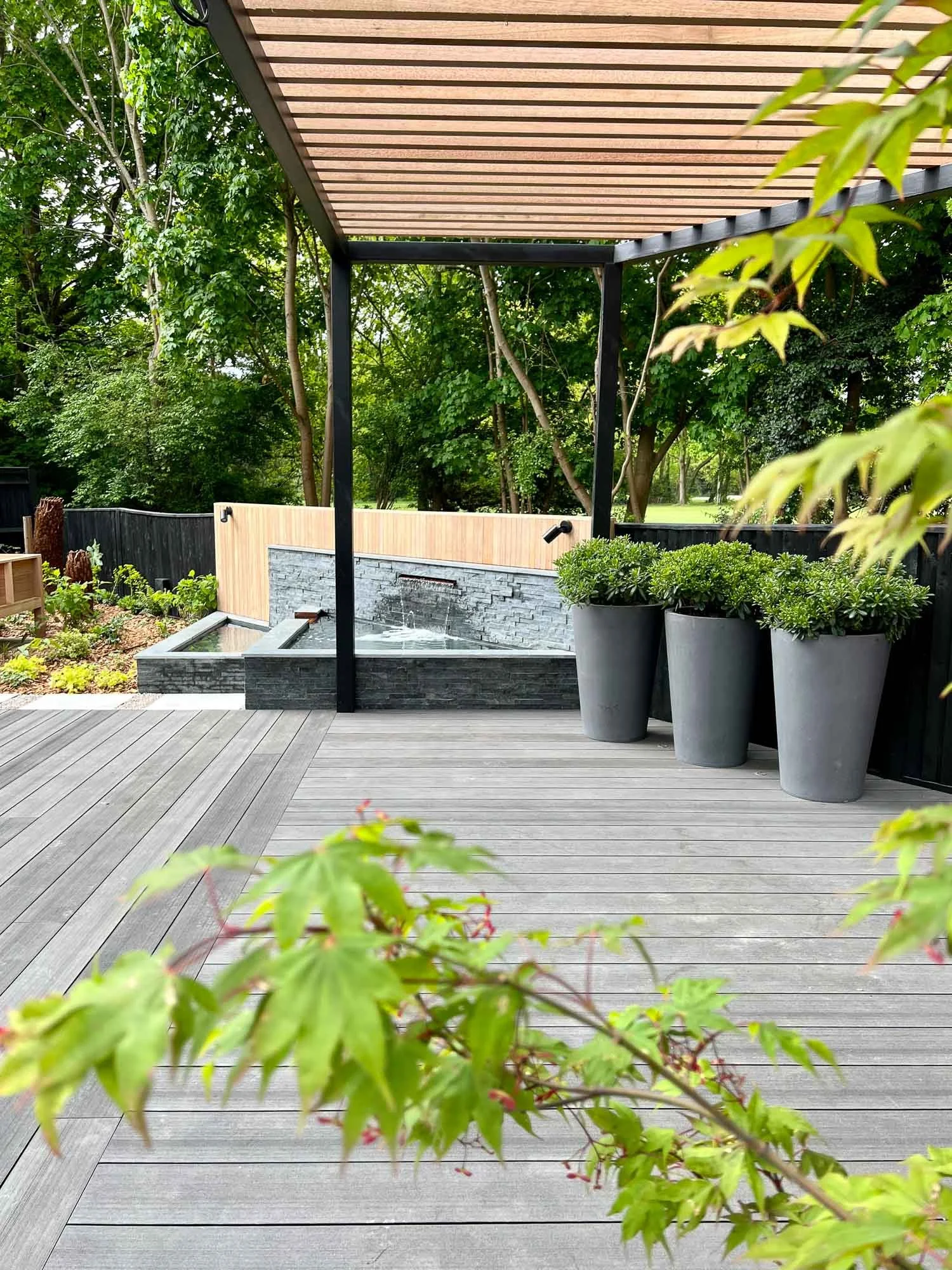 Modern patio with wooden decking, a small water feature with stone walls, large gray planters with greenery, and a wooden trellis overhead, surrounded by lush green trees and a black fence.