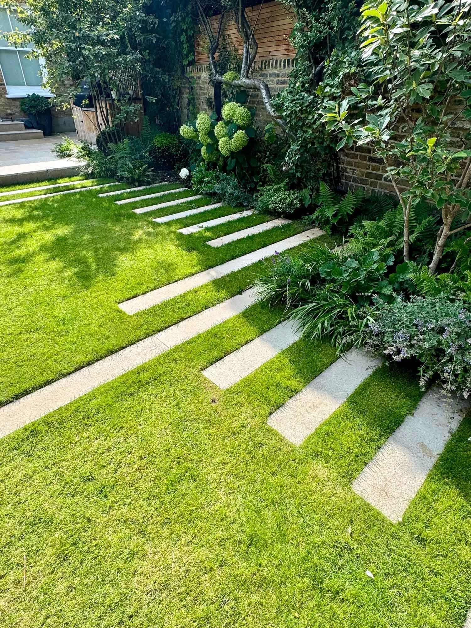 Garden with a striped stepping stone path, lush green grass, and various shrubs and trees along a brick wall.