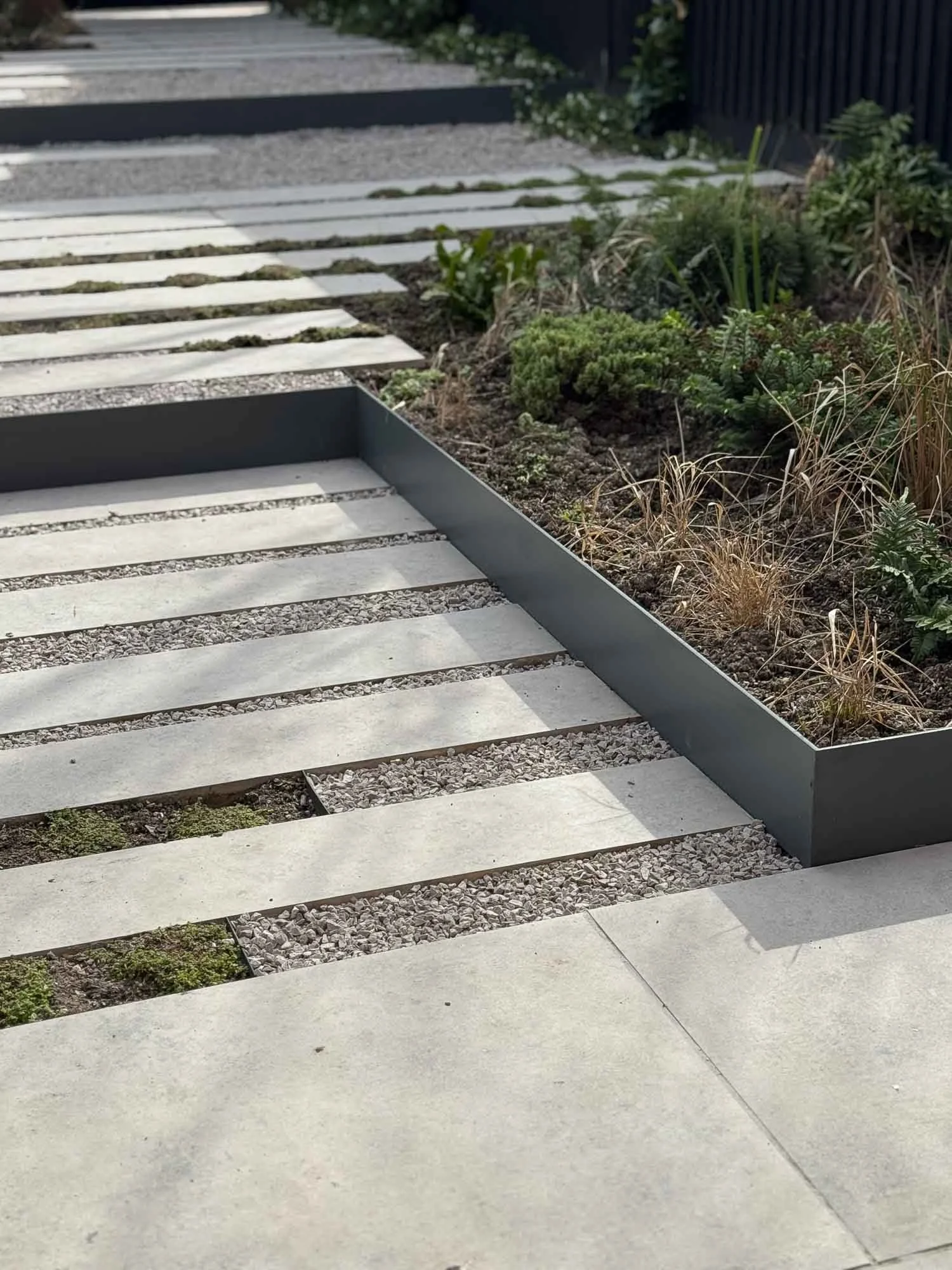Close-up of a modern garden pathway with rectangular white concrete stepping stones separated by small gravel and a raised garden bed with various plants on the side.