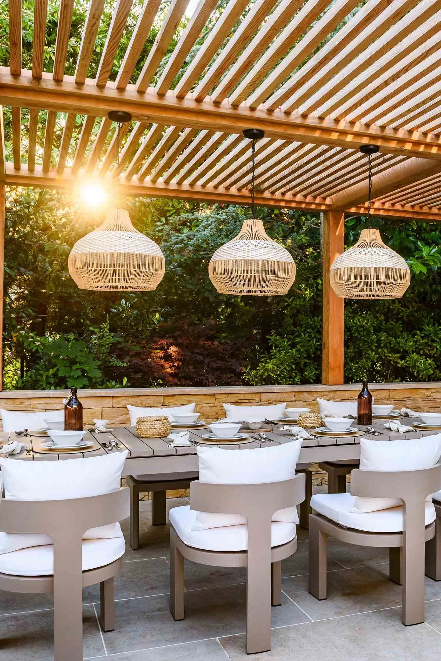 Large outdoor dining table under a timber pergola with three woven pendant lights and white upholstered chairs