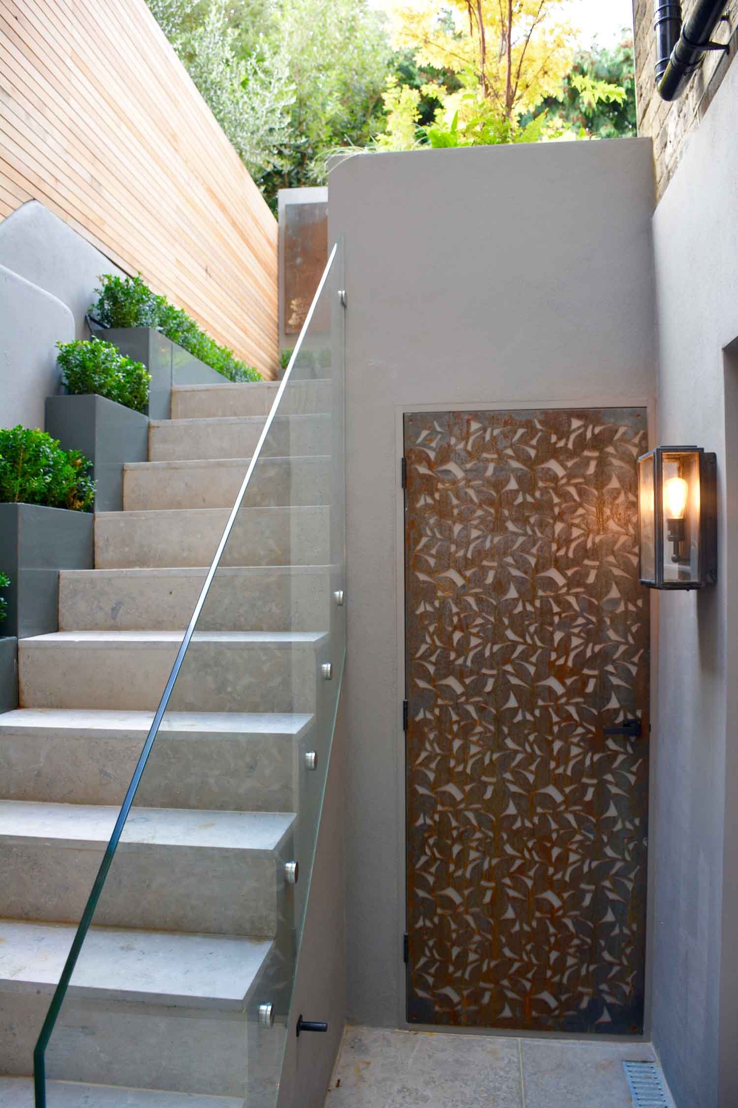 Outdoor staircase with glass railing, leading up alongside potted plants, with a decorative door and a wall-mounted light fixture beside it.