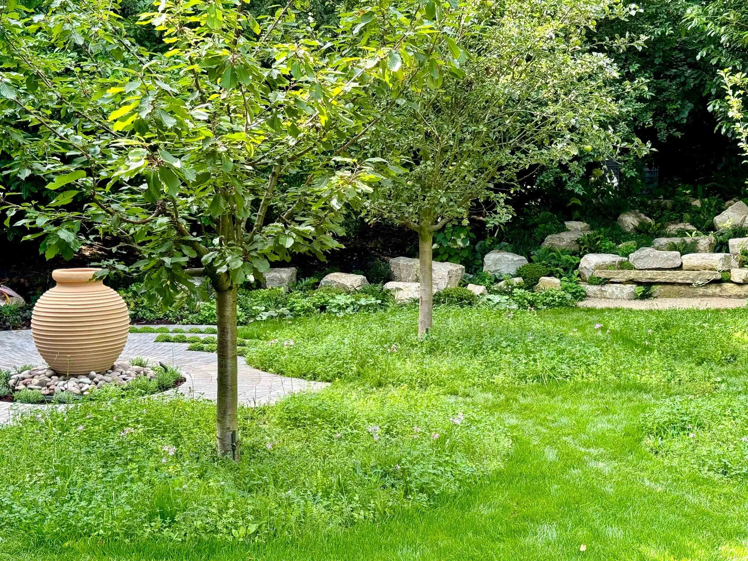 A lush green backyard with two trees, a decorative beige vase on a small paved area, and a stone garden border.