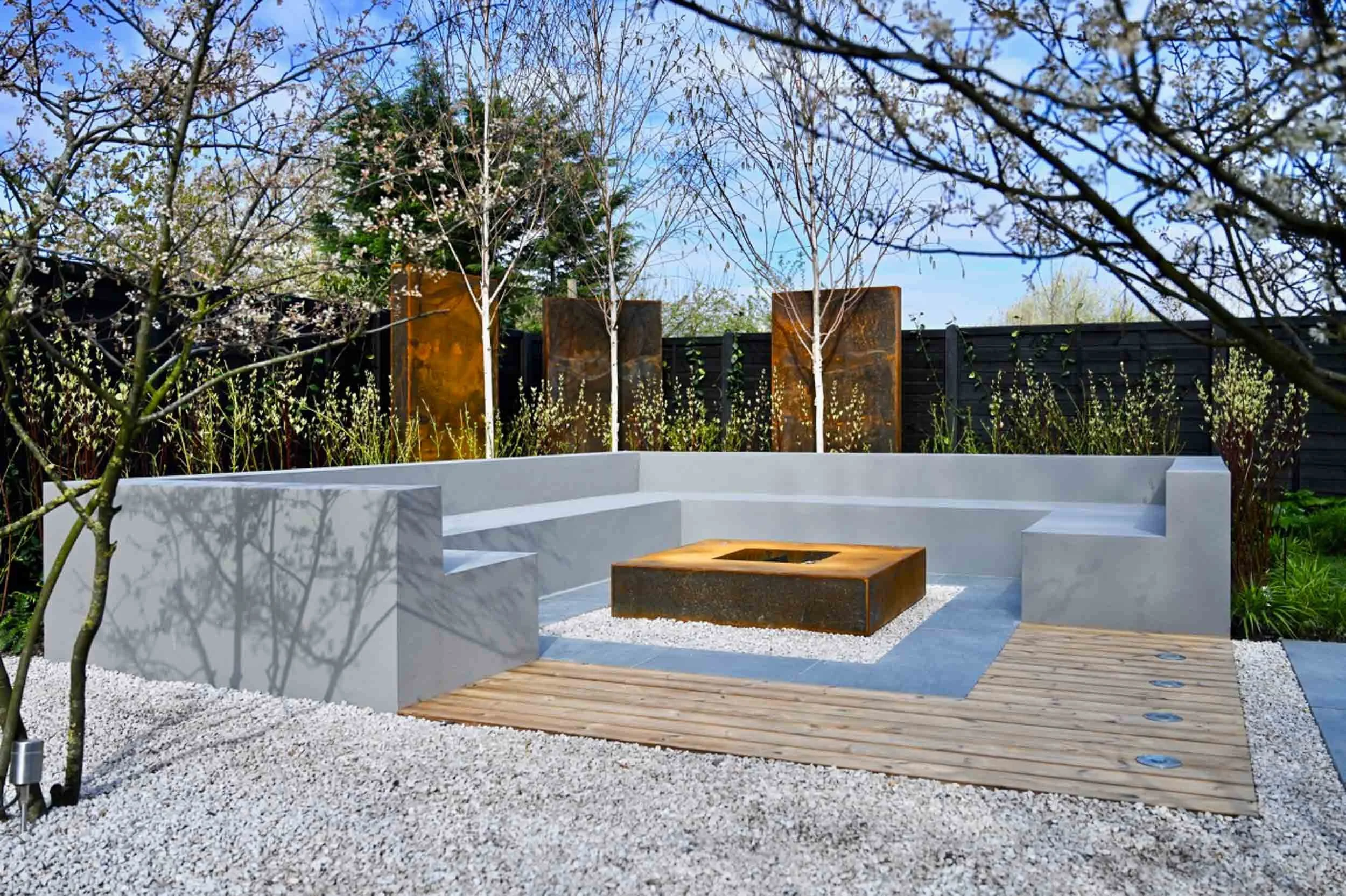 Modern outdoor seating area with white concrete benches, wooden decking, gravel, and a small fire pit in the center. Surrounding trees and plants, black fence, and garden sculptures in the background.