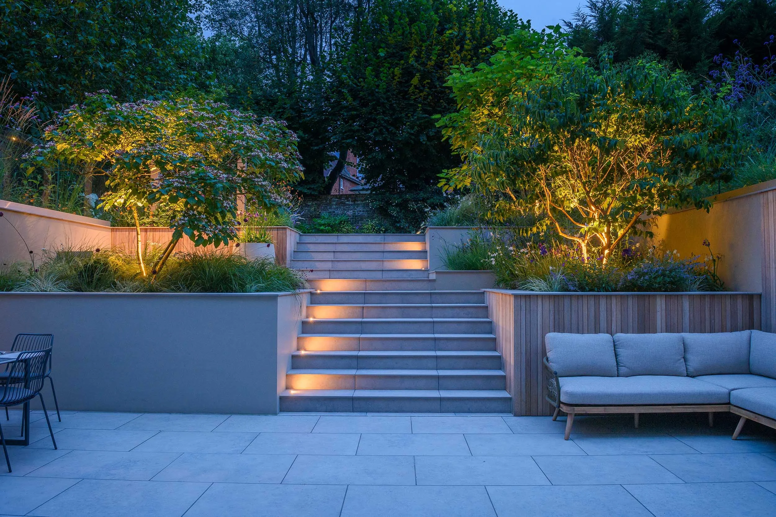 Evening view of illuminated garden steps and uplighted trees creating a warm glow across a modern paved terrace area