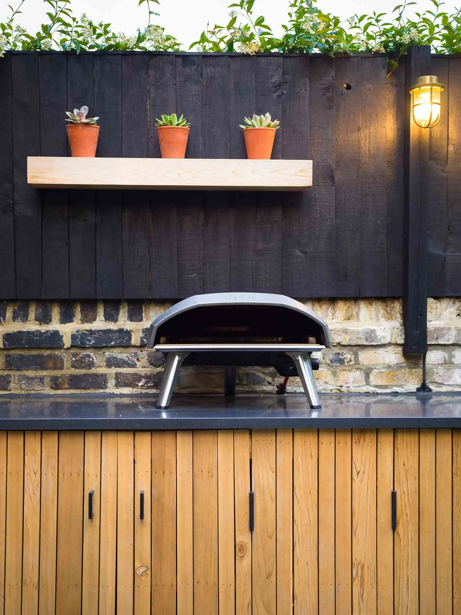 Modern black pizza oven on a wooden countertop with small succulents in terracotta pots on a shelf