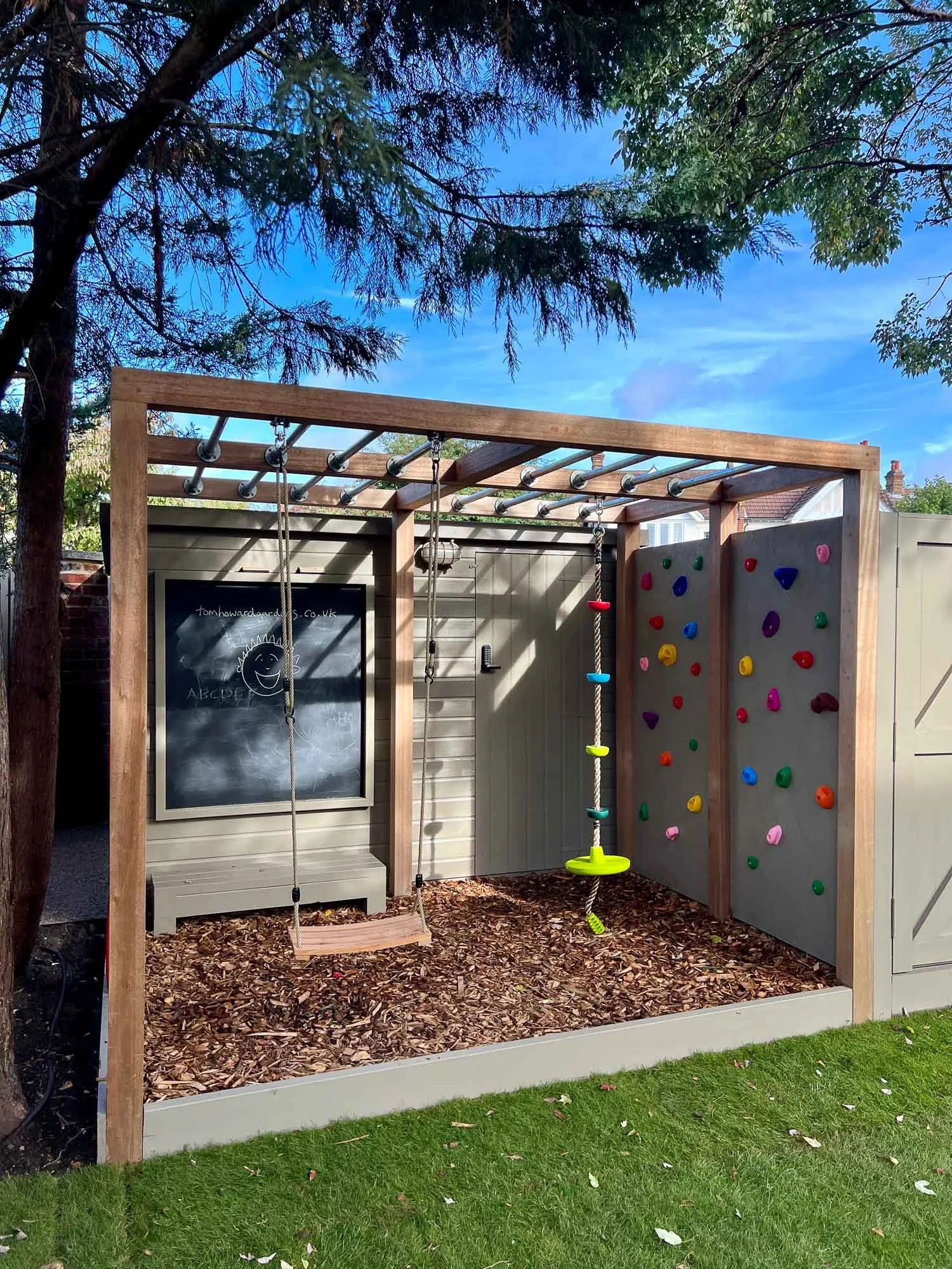 Wooden outdoor play structure with swings, a climbing wall with colorful holds, a rope ladder, and a chalkboard wall, situated on wood chips with grass in the foreground and trees and a blue sky in the background.