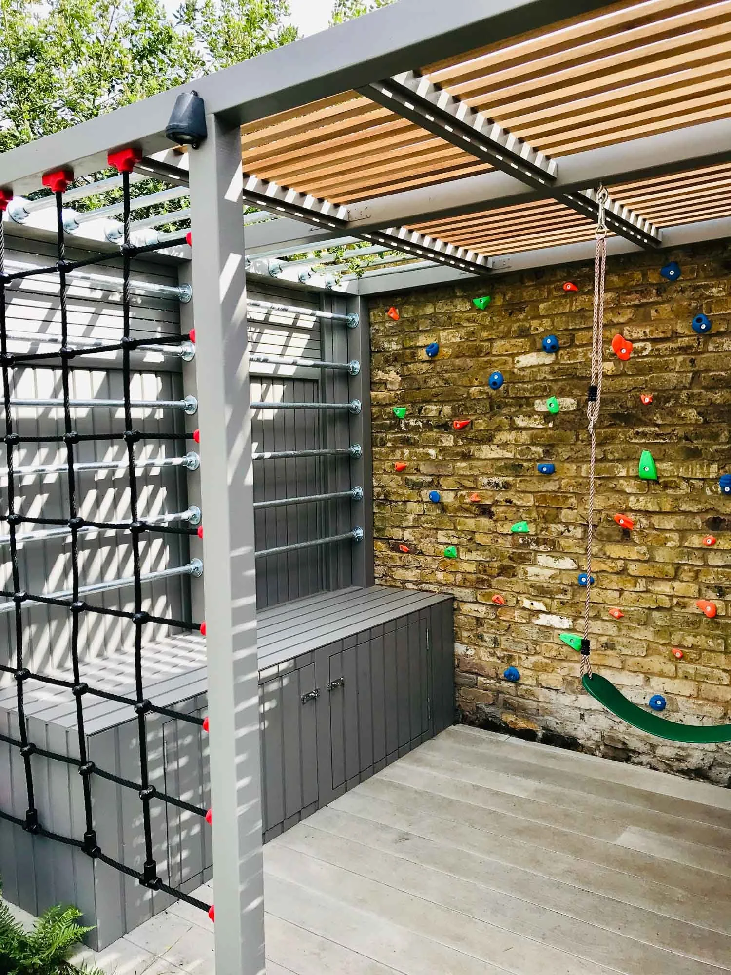 Outdoor play area with climbing wall, rope swing, and shaded wooden roof, enclosed by brick wall and metal fencing.