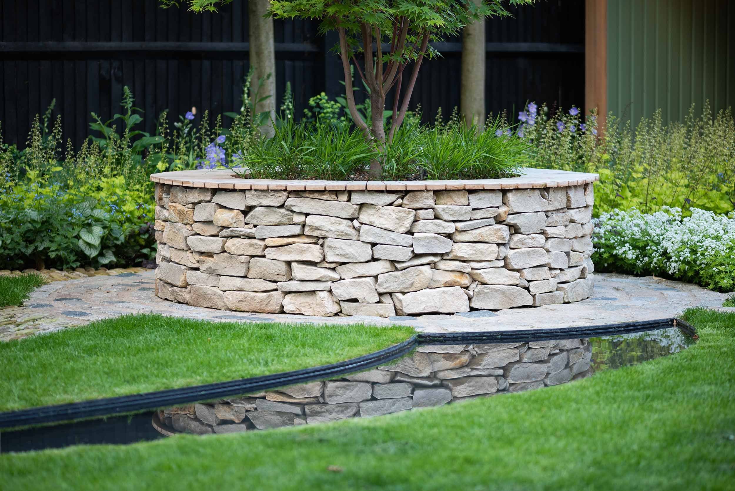 A circular stone garden bed with a young tree and green plants surrounded by a multicolored stone pathway and lush grass.