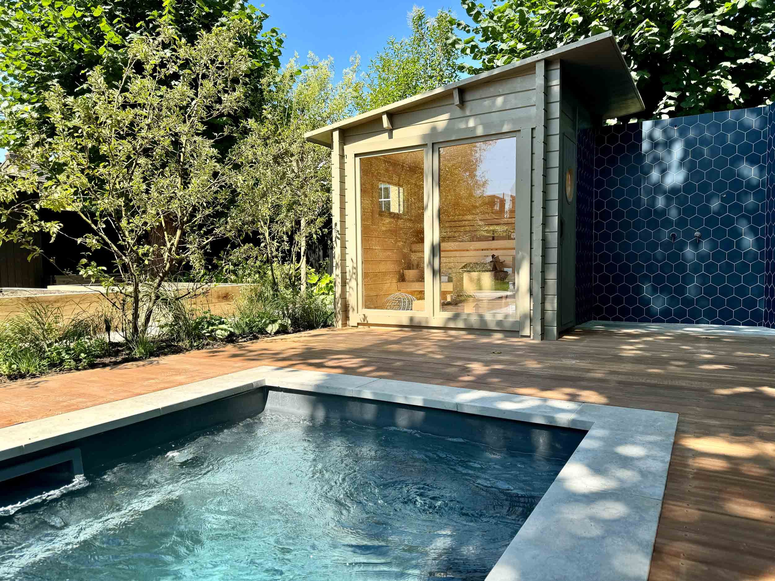 A backyard with a small hot tub, wooden deck, lush green trees, and a wooden structure with a glass window, set against a bright blue sky.