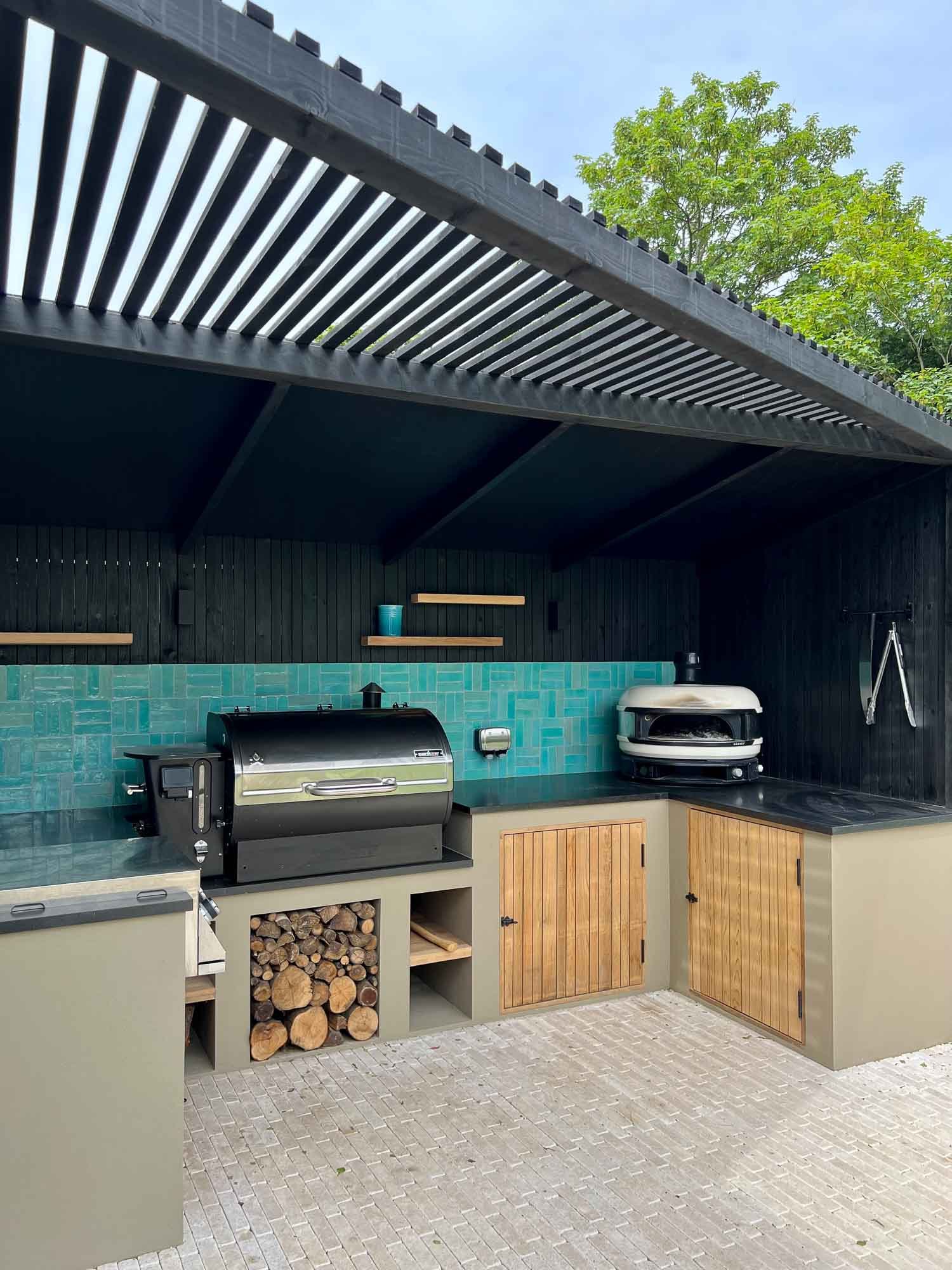 Outdoor kitchen with two grills, wood storage, and wooden cabinets, under a black pergola with green trees in the background.