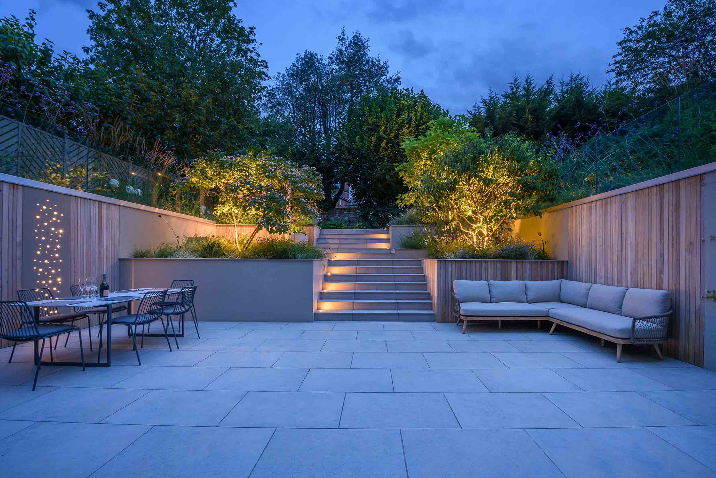 Evening view of a large stone patio with an illuminated garden staircase and a contemporary wooden corner sofa