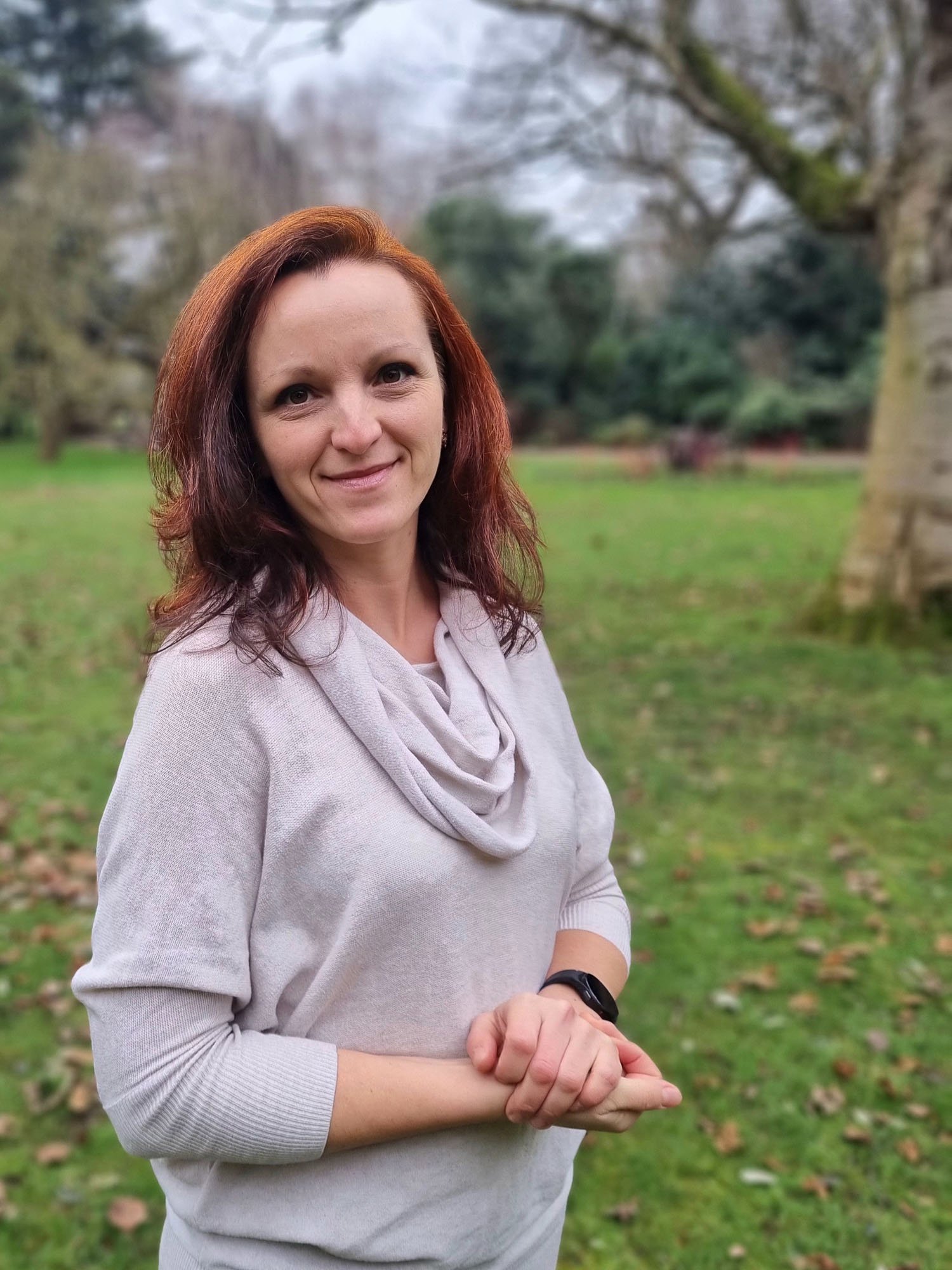 Smiling woman with long reddish hair wearing a light cowl neck jumper standing in a green park with trees in the background