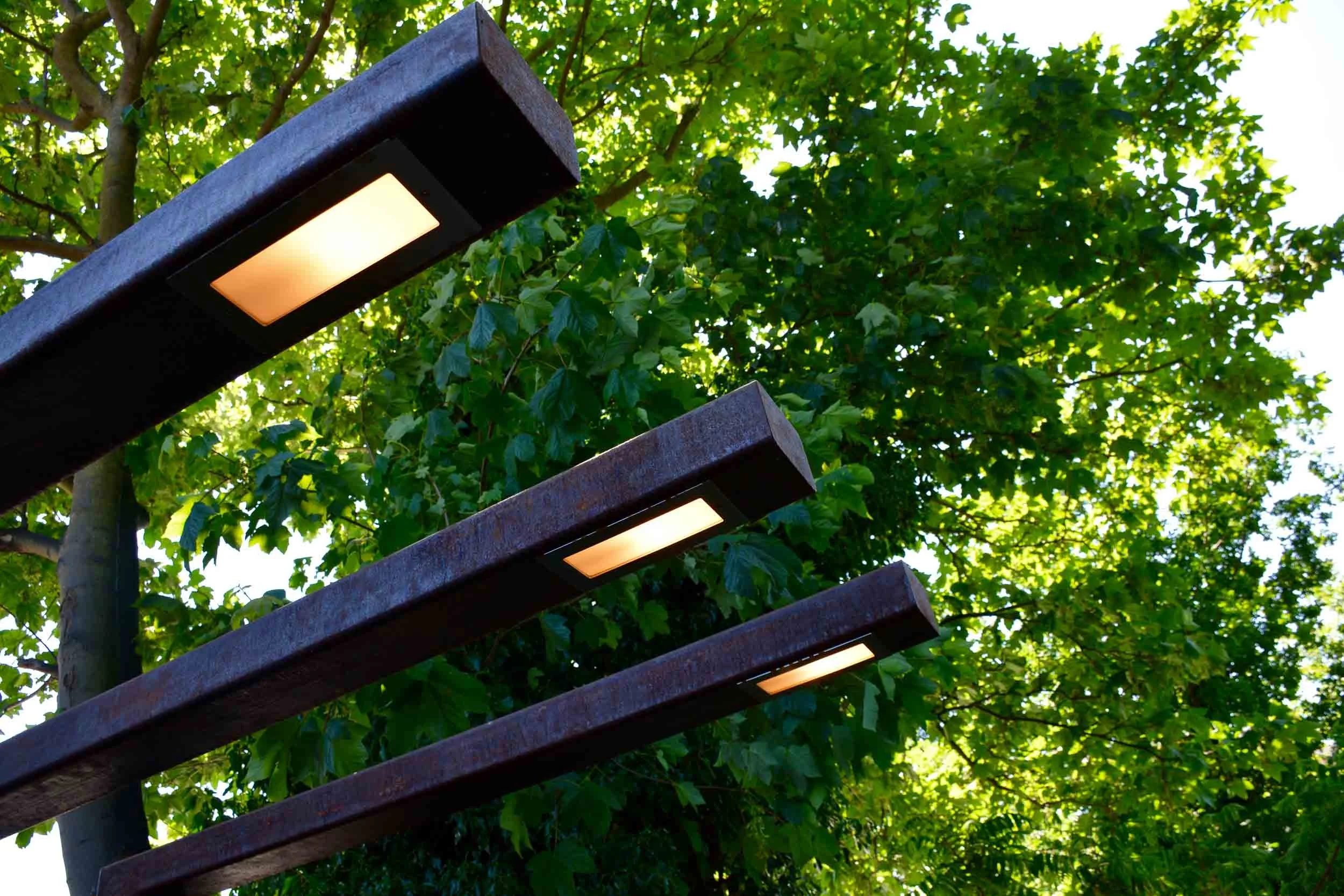 Outdoor modern light fixtures mounted on a dark metal structure with green leafy trees in the background.