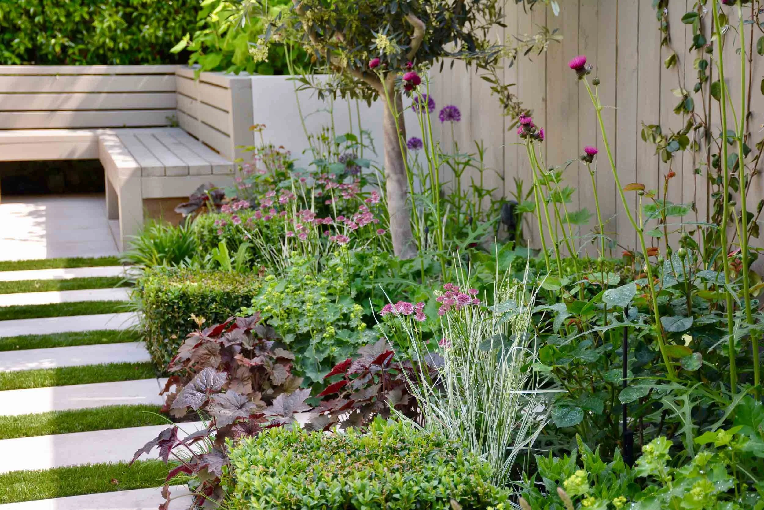 A lush garden with various flowering plants, shrubs, and tall purple and pink flowers next to a wooden fence and a white bench in the background.