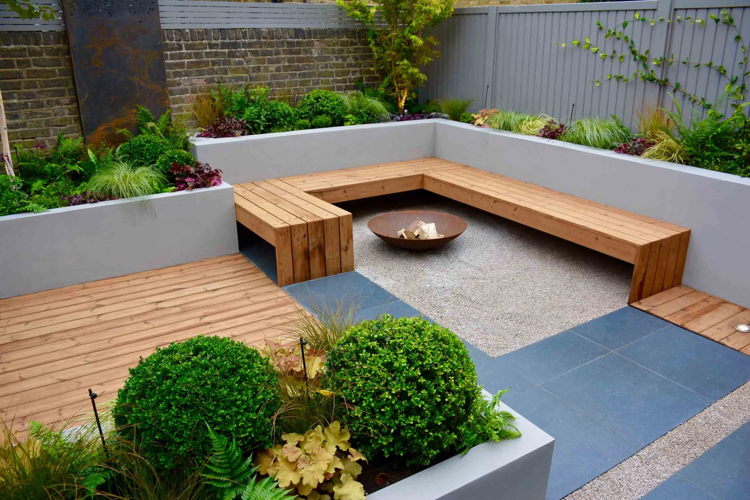 Outdoor garden patio with a built-in wooden bench, concrete borders, a fire pit, and lush plants along a gray fence and brick wall.