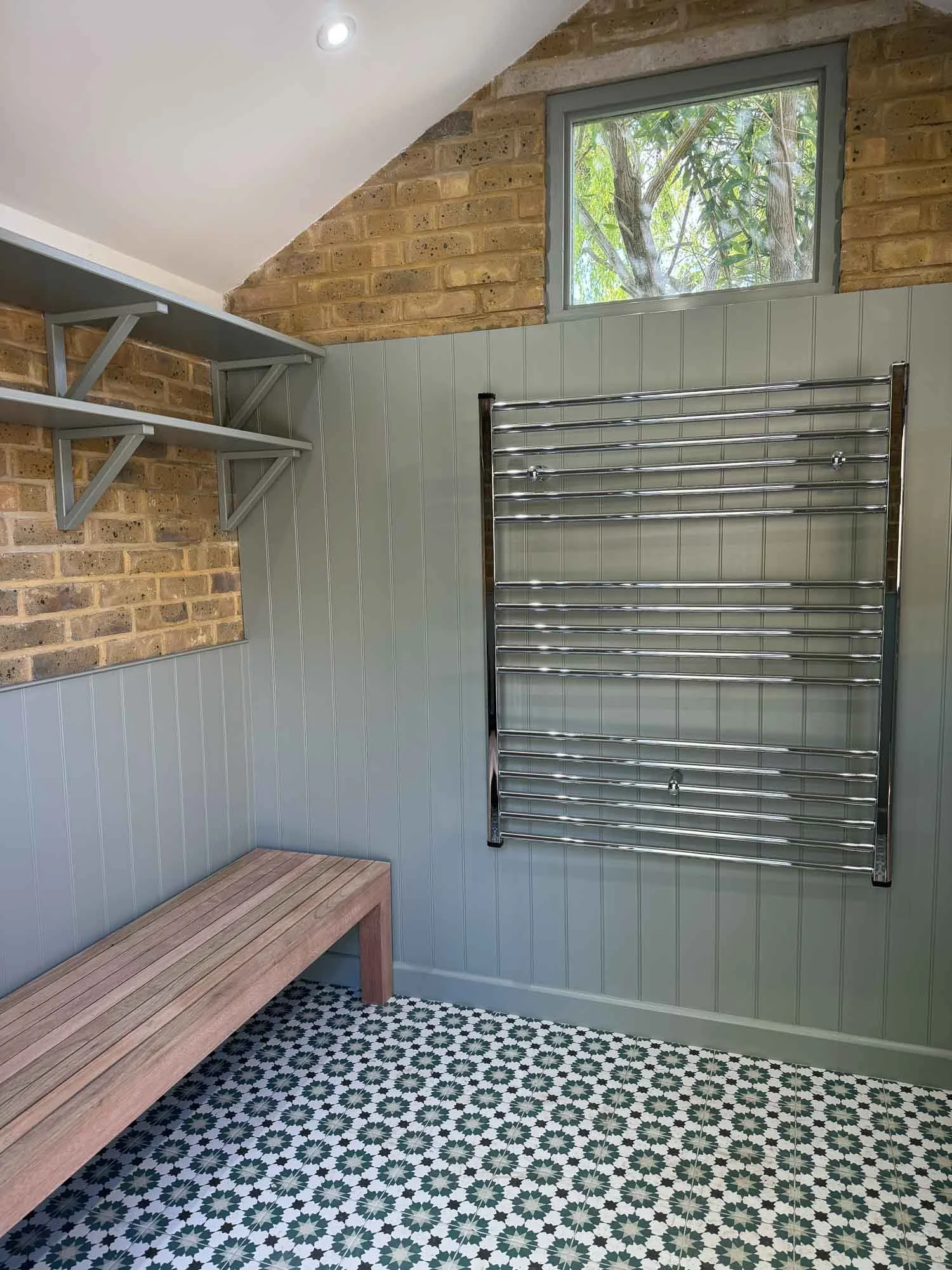 A cozy room with mixed wall materials including brick and beadboard paneling. There is a small window near the ceiling, allowing natural light in. The room features a set of gray shelves mounted on the brick wall, a sleek silver towel rack mounted on