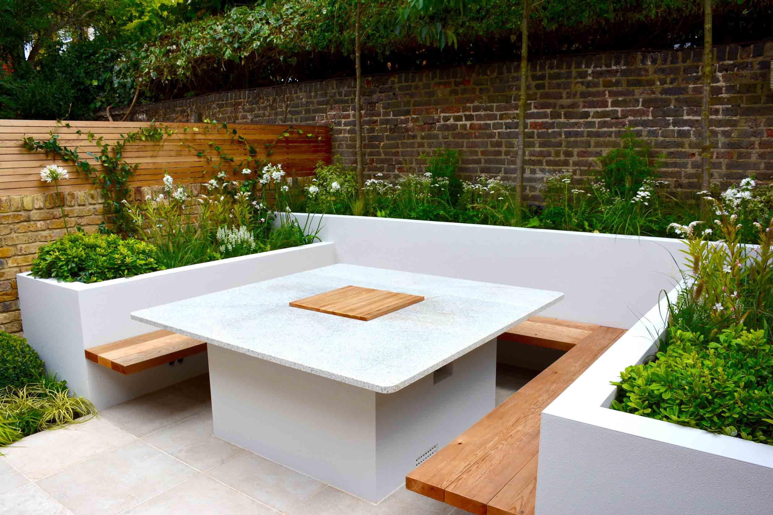 Outdoor patio area with built-in white concrete seating, wooden benches, a square table with a wooden centerpiece, and surrounding lush green plants against a brick wall.