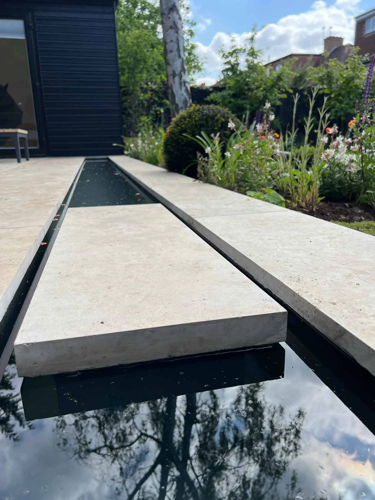 A modern outdoor fountain with rectangular stone slabs surrounding a narrow water channel, reflecting trees and the sky, with a garden and house in the background.