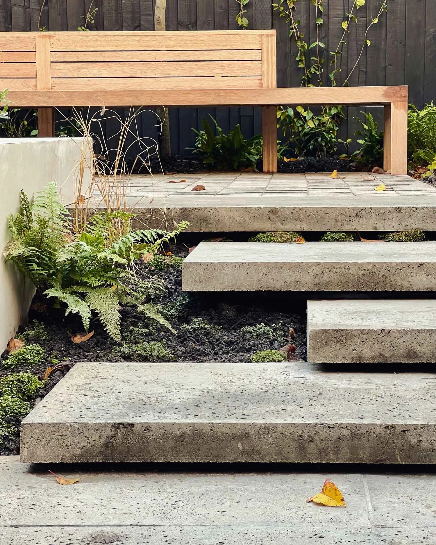 Concrete stepping stones leading to a wooden bench in a garden with plants and a dark wooden fence.